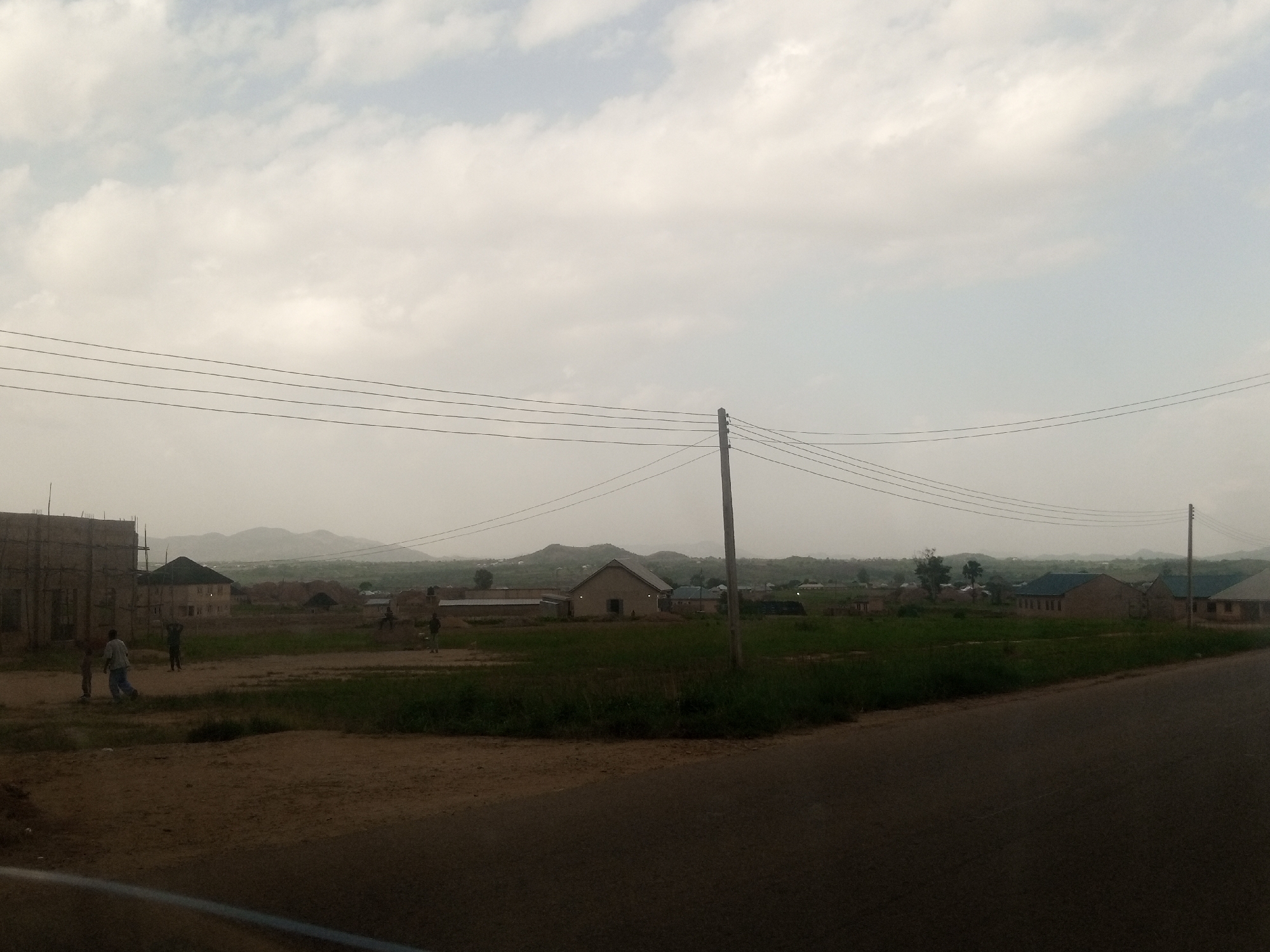 The image shows a rural or semi-urban landscape under a cloudy sky. In the foreground, a dirt road curves into the right side of the frame. Power lines stretch horizontally across the top half of the image, supported by a utility pole visible on the right.
The midground features several buildings, some appearing to be under construction, surrounded by green grass and dirt patches. A few people are scattered throughout this area, with some gathered near a partially constructed building on the left. The figures are small and their activities are not clearly discernible. In the distance, a cluster of homes with light-colored walls and darker roofs are visible, suggesting a small settlement. Further back, rolling hills or mountains form the backdrop, softened by atmospheric haze.
The time of day appears to be overcast, possibly late afternoon or early morning, given the diffused light and muted colors. The weather seems to be calm, with no strong indications of rain. The overall atmosphere is quiet and ordinary, depicting a scene of everyday life in a developing area. There is no visible text in the image.