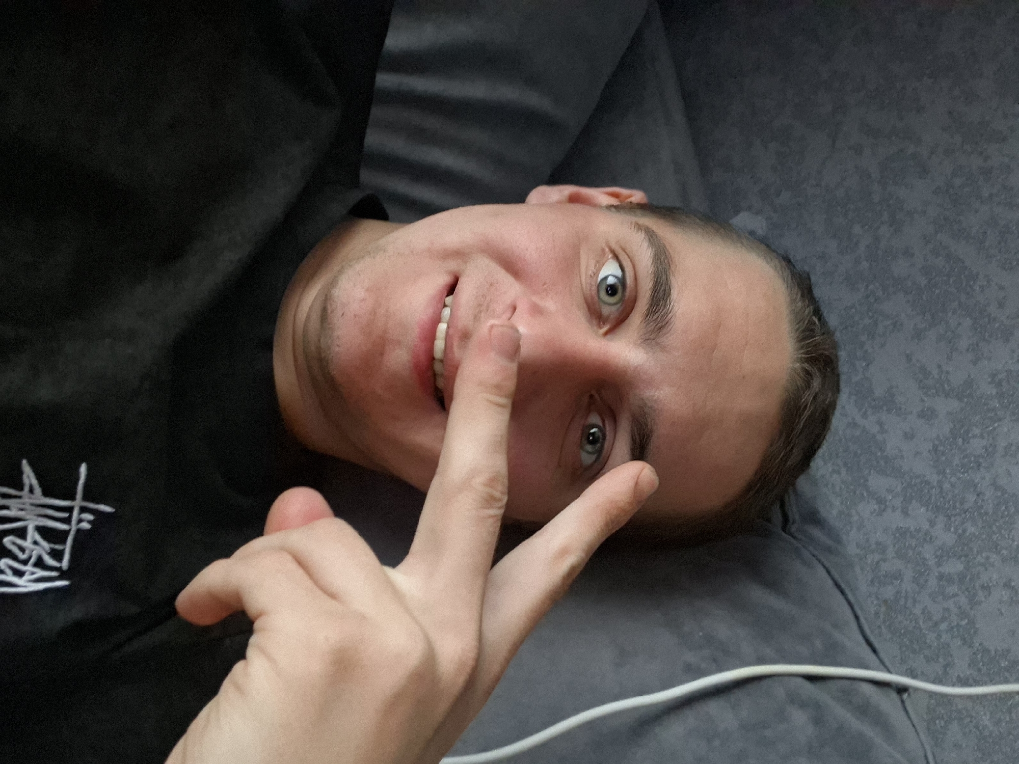 A man is lying on a grey couch, facing the camera, with his right hand raised and two fingers touching his nose. The man is wearing a black shirt with a white design on the left chest. His hair is short and light brown. The background is out of focus, but appears to be a couch and possibly a wall. There is a white cable visible on the couch. The location and city are unknown.