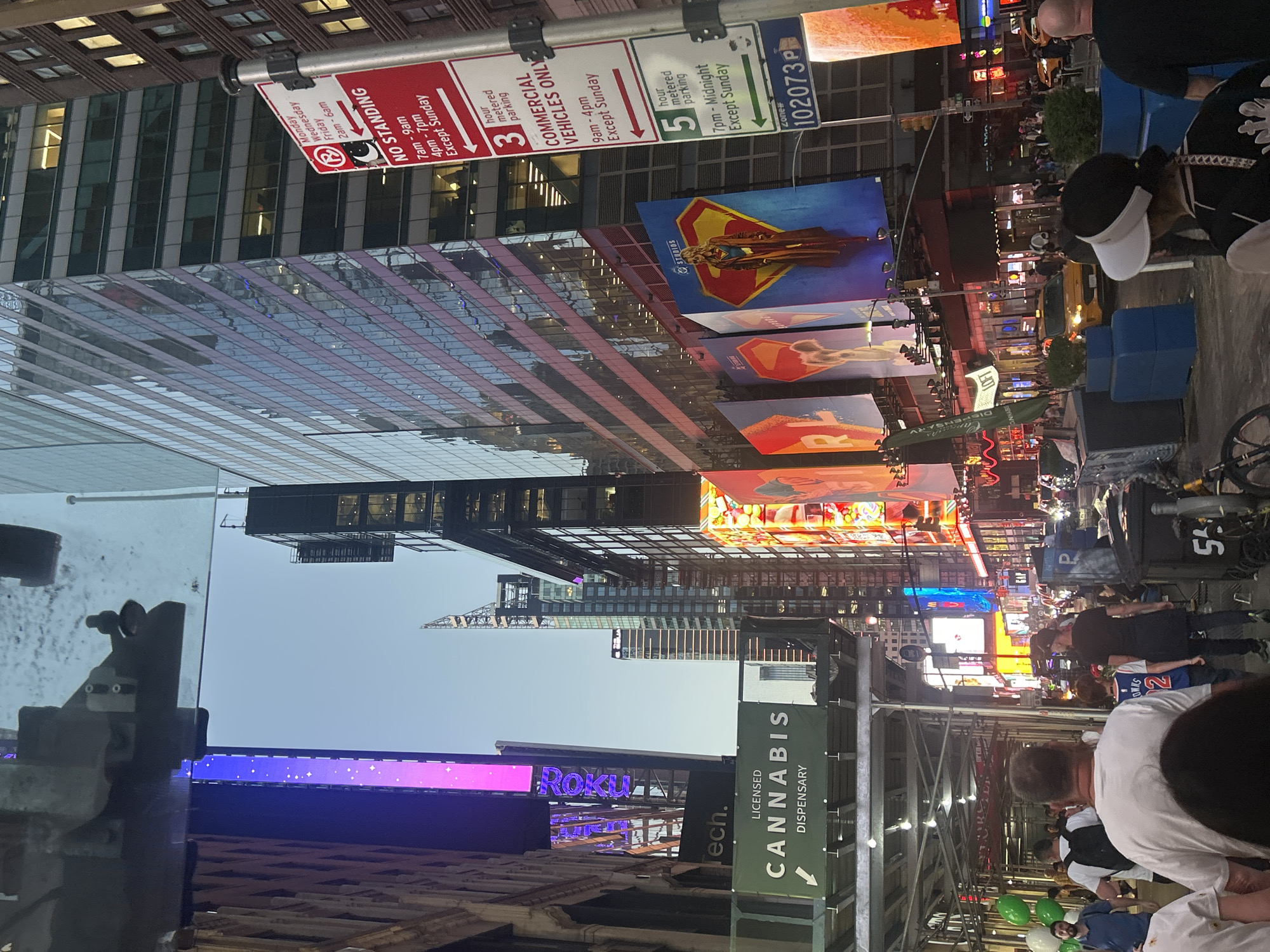The image captures a vibrant, bustling urban street scene at night or dusk, characteristic of a major city like New York City. Towering skyscrapers with reflective glass facades line the street, their surfaces reflecting the myriad of bright, colorful digital billboards and neon lights that dominate the midground and background.
Numerous people populate the scene, primarily visible in the lower right portion, suggesting a lively pedestrian area. They appear to be walking and congregating, though no specific event is discernible beyond general city life. In the foreground, a person wearing a white visor and another in a white shirt are visible from behind. A child in a blue jersey with the number "2" on it is also present.
The street is adorned with multiple informational signs. A red "NO STANDING" sign specifies restrictions for Monday, Wednesday, and Friday from 2 am-6 am, 7 am-9 am, and 4 pm-7 pm, with an exception for Sunday. Adjacent white and green signs indicate "3 hour metered parking" for "COMMERCIAL VEHICLES ONLY" from 9 am-4 pm, except Sunday, and "5 hour metered parking" from 7 pm-Midnight, except Sunday. A "Zone" number "102073 P" is also visible.
Prominent digital billboards display various advertisements. One large billboard features a figure resembling Superman or Supergirl, next to the word "STUDIOS." Another screen brightly displays the "Roku" logo. At street level, a large green sign points downwards, reading "LICENSED CANNABIS DISPENSARY," indicating a specific type of business. A yellow taxi cab is partially visible in the mid-right amidst the bright lights. Construction scaffolding is present in the lower foreground. The overall impression is one of a dynamic and commercially active city environment after dark.