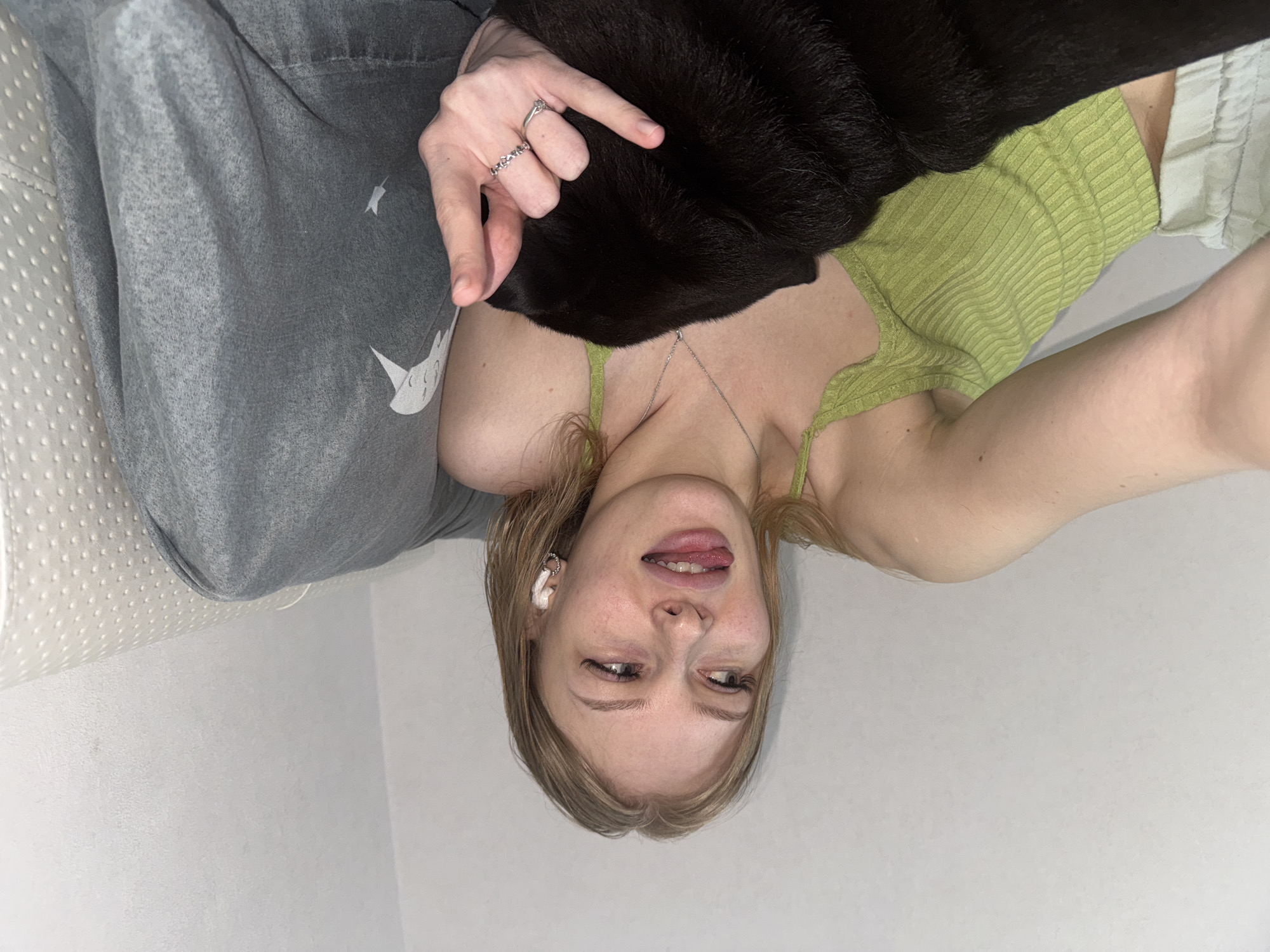 The media file is a self-portrait taken indoors. A young woman with blonde hair is lying down, holding a black cat. She is wearing a green ribbed tank top and grey sweatpants with a white logo. The woman is sticking her tongue out, and her eyes are partially closed, suggesting a playful or relaxed mood. She is wearing small hoop earrings and several rings on her finger. She also has white earbuds in her ears. The background appears to be a light-colored wall, and there is a textured surface on the left side of the image, possibly a couch or bed. The lighting suggests it is daytime, and the setting is an indoor environment. There is no visible text in the image.