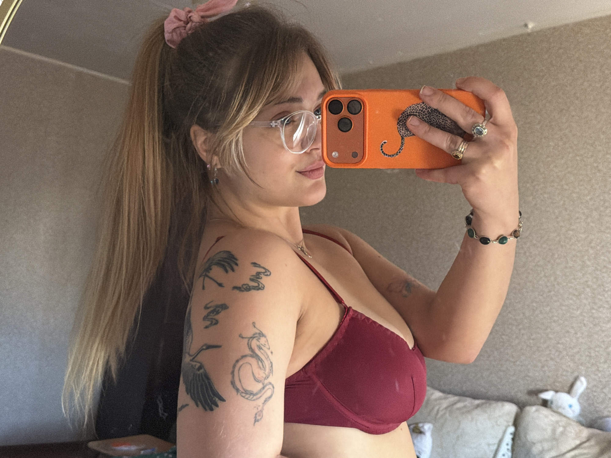 A young woman is holding an orange cell phone, with a drawing of a leopard on the back, and taking a selfie. She is wearing clear framed glasses, a pink scrunchie in her hair, a burgundy bra, and several pieces of jewelry, including rings and a bracelet. She has several tattoos on her left arm. The location is a room with a neutral colored wall and a couch with pillows visible in the background. There is no indication the photo was taken in Naberezhnye Chelny, Russia.