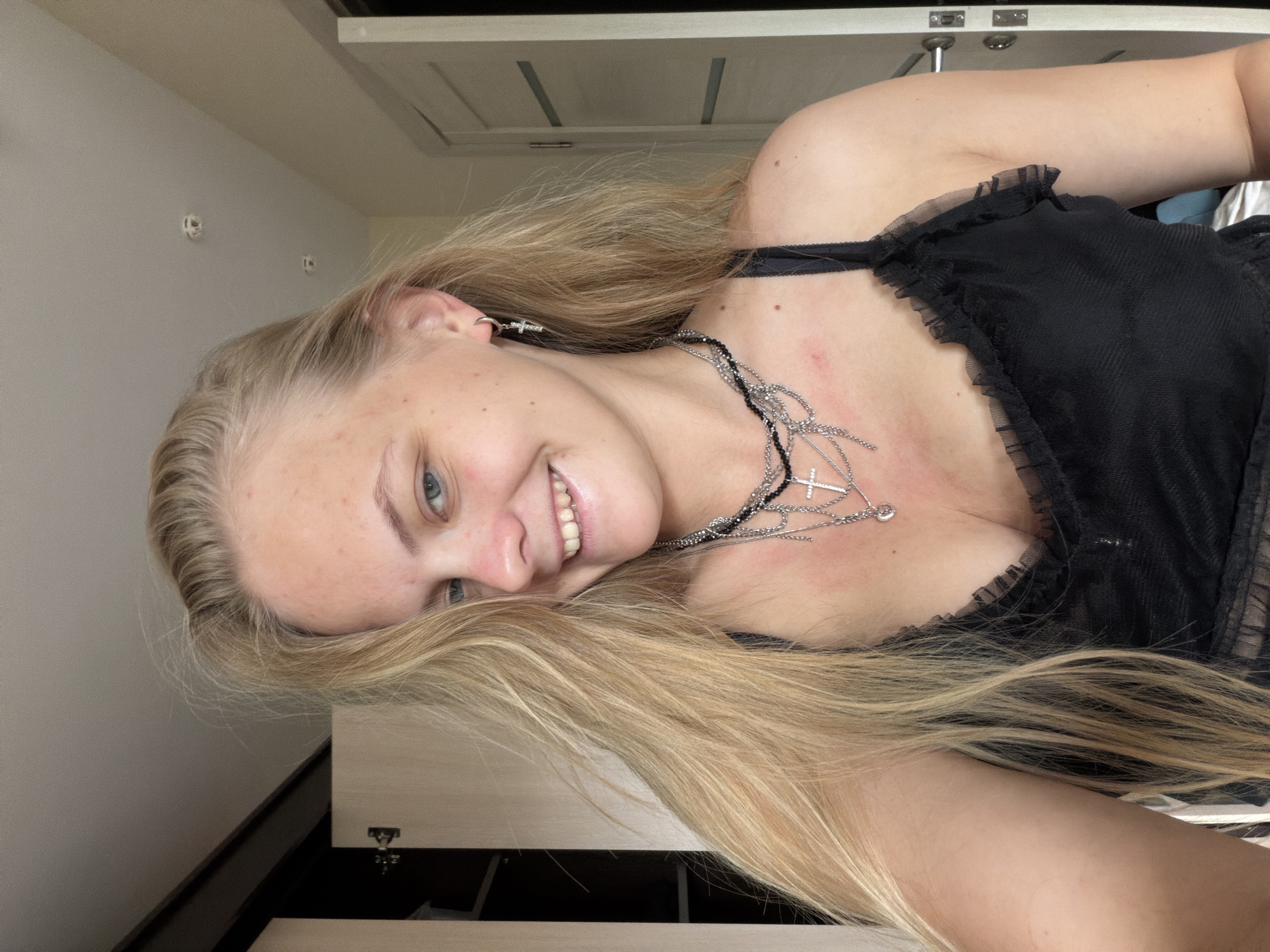 A young woman with long, straight blonde hair and blue eyes is captured in a close-up shot, rotated 90 degrees counter-clockwise. She is smiling broadly, showing her teeth, and looking directly at the camera. Her complexion is fair, with some minor reddish blemishes or freckles visible on her forehead, cheek, and chest.
She is wearing a black top or dress that features a sheer, ruffled fabric around the neckline and shoulders, possibly made of tulle. Adorning her neck is a prominent, multi-layered silver-toned necklace with several chains and at least two cross pendants, one of which is larger and more intricate. Matching silver-toned cross earrings are visible in her left ear.
The setting appears to be indoors. Behind her head and shoulders, a plain light-colored wall is visible, with two small, indistinct white objects on it. The upper background features a light-colored surface with horizontal dark lines, possibly part of a door, cabinet, or ceiling panels; hinges are discernible on this surface in one crop. Below her, there's a darker area with a lighter-colored, possibly wooden or laminate, surface, suggesting furniture like a dresser or desk. The lighting is soft and even. No visible text is present in the image. The woman conveys a happy and friendly emotion through her smile.