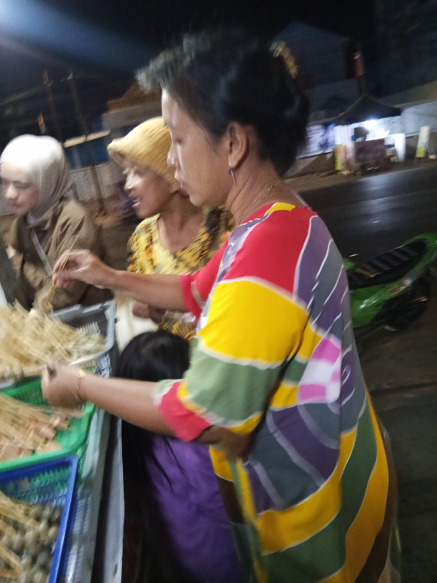 The image depicts a street scene at night in Lubuklinggau, Indonesia, likely a market or roadside stall. In the foreground, a woman in a brightly colored, patterned tunic with yellow, red, purple, and green sections is interacting with a display of food on skewers. Her right hand is extended towards the skewers, suggesting she is either preparing them or serving them. To her left, partially visible, is another woman wearing a yellow head covering and a yellow, patterned top, and behind her, another individual with white head covering and a brown garment. A child, possibly, is visible between the two women, wearing a purple garment. The background shows a street with a motorcycle and the blurred outlines of buildings or stalls, illuminated by artificial light, suggesting a bustling evening atmosphere.