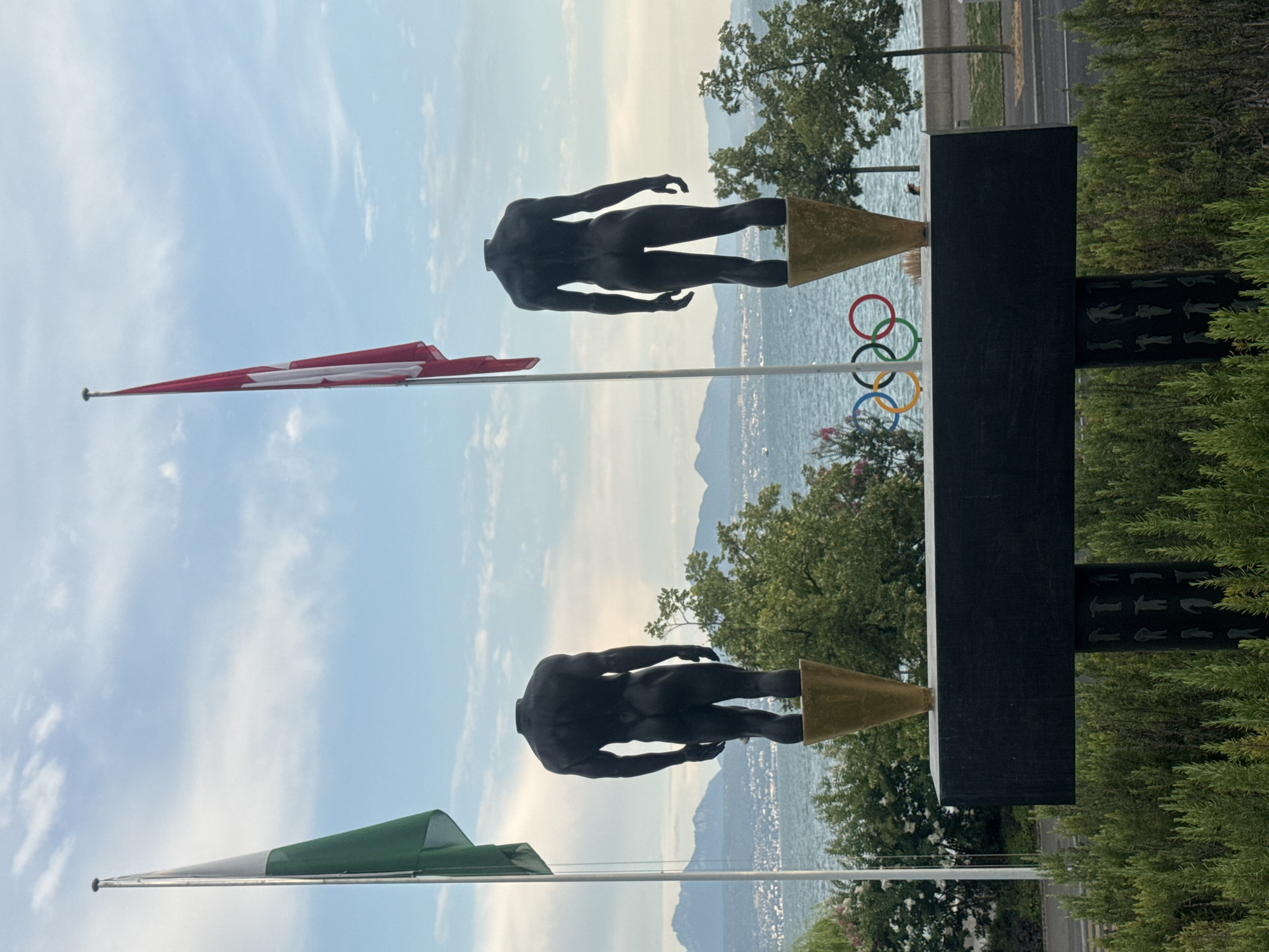 The image depicts two headless male statues standing on golden conical bases, positioned behind a black rectangular plinth. The statues are dark in color and appear to be made of bronze or a similar material. They are facing away from the viewer, towards a body of water with mountains in the distance.

To the left of the statues, a green and white flag on a flagpole is visible, and to the right, a Swiss flag (red with a white cross) is also on a flagpole. Directly in front of the plinth, the Olympic rings are displayed. The background features lush green trees and bushes, a calm body of water, and a distant cityscape nestled against a backdrop of mountains. The sky is cloudy with patches of blue.

This scene is located at the Olympic Museum in Lausanne, Switzerland. The Olympic Museum is situated on the shores of Lake Geneva, with the French Alps visible across the water in the background. The statues are part of a sculpture display outside the museum.