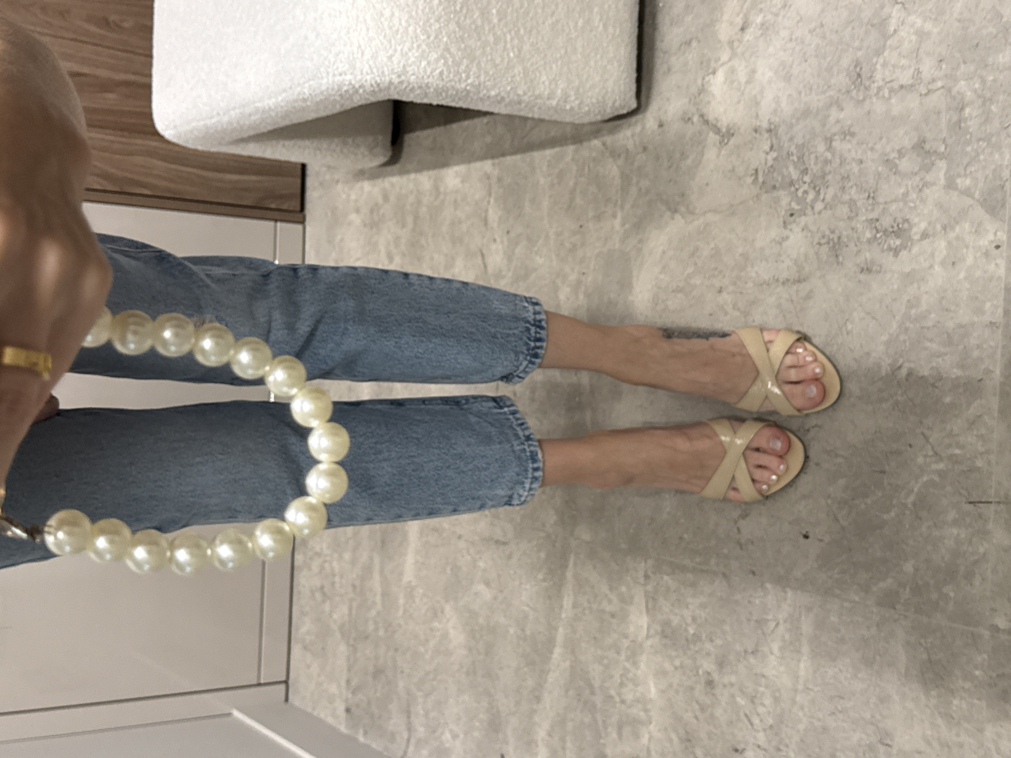 A person is standing on a marble floor in what appears to be an indoor setting, possibly a home or a boutique. The person is wearing light-wash denim jeans and light beige slide sandals with criss-cross straps. They are holding a large pearl necklace. The background includes a white textured chair or bench and part of a wooden wall or door frame. There is no information in the image to suggest the location is Moscow, Russia.