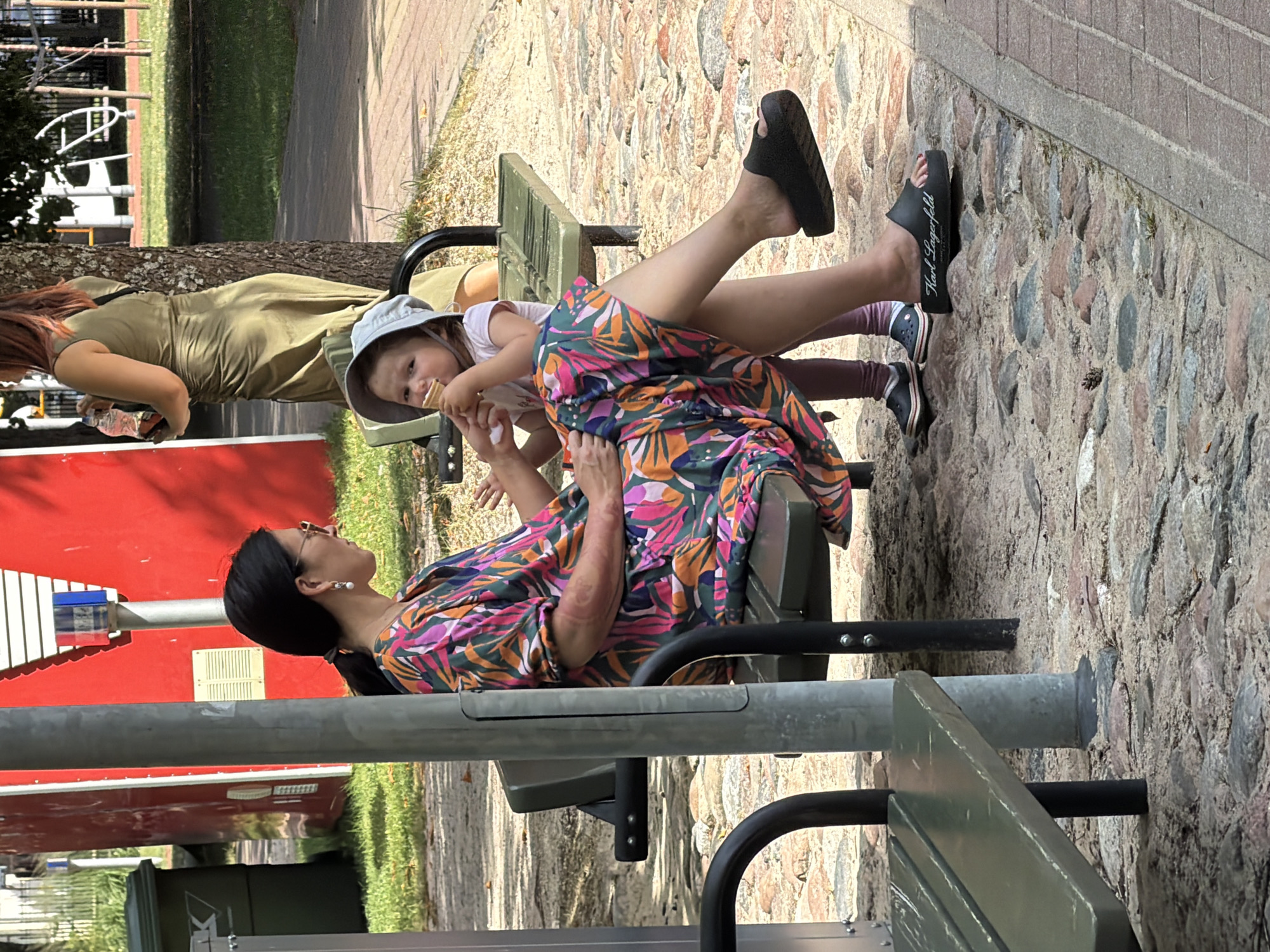 In Ventspils, Latvia, on a sunny day, a woman wearing a vibrant, colorful dress and sunglasses is seated on a park bench. She is accompanied by a young child, also seated on the bench, who is wearing a light-colored hat and eating an ice cream cone. To the right of the bench, another woman in a green dress stands holding a bottle of water. The scene is set outdoors, with a paved walkway in the foreground and a grassy area with trees in the background. The bench appears to be part of a public park or recreational area, indicated by the presence of other seating and playground equipment visible in the distance. The ground surface is a mixture of sand and cobblestones.
