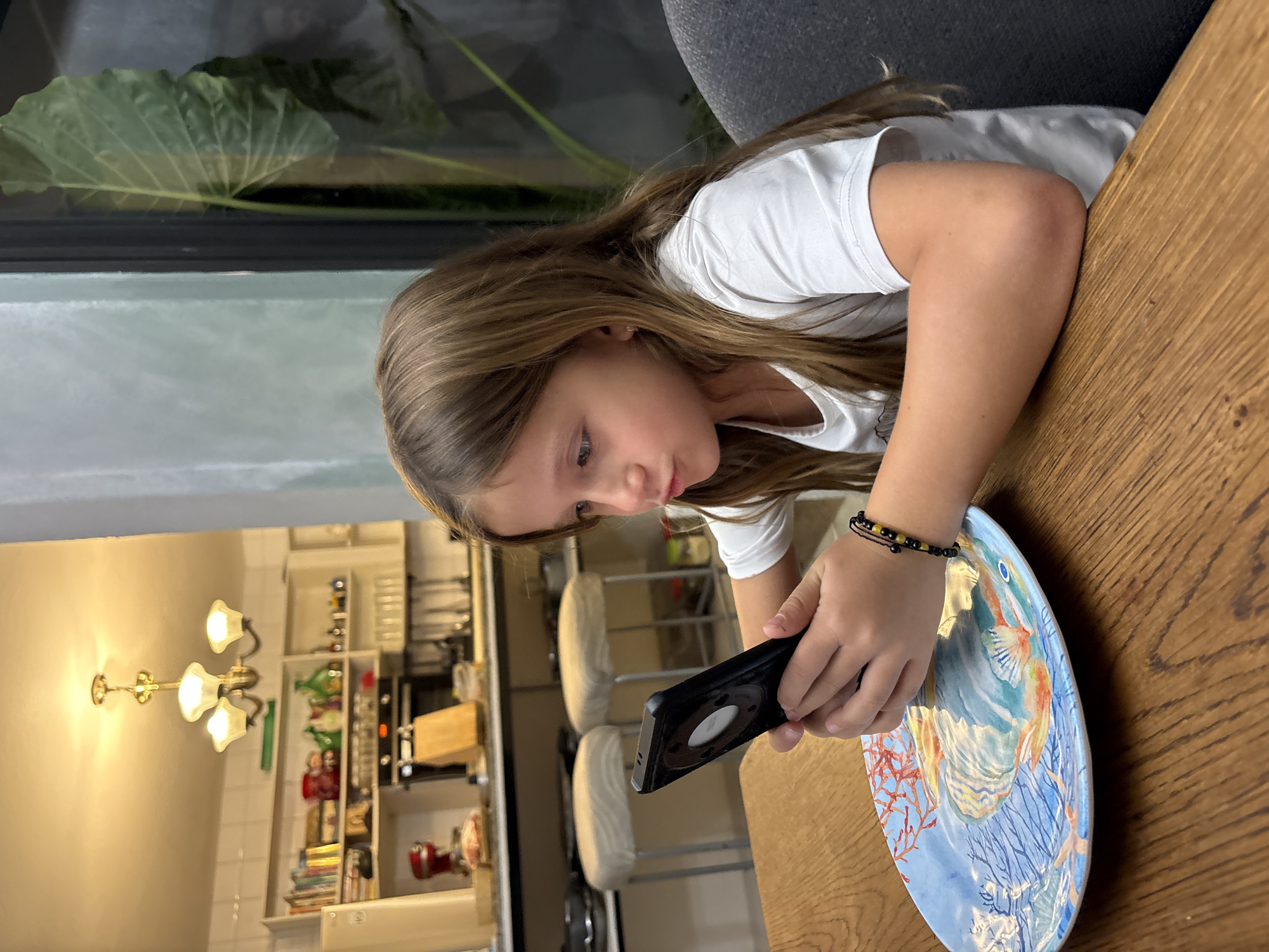 A young girl with long brown hair is seated at a wooden table, intently focused on a black smartphone held in her hands. She is wearing a white t-shirt and a beaded bracelet on her wrist. Next to her is a decorative plate with a blue and orange design featuring fish and coral. In the background, a kitchen area is visible, including a wall-mounted light fixture, shelves stocked with dishes and appliances, and chairs. The scene appears to be indoors, with natural light filtering through a window or glass door. The location is not ascertainable as Puebla, Mexico, based on the visual content of the image.