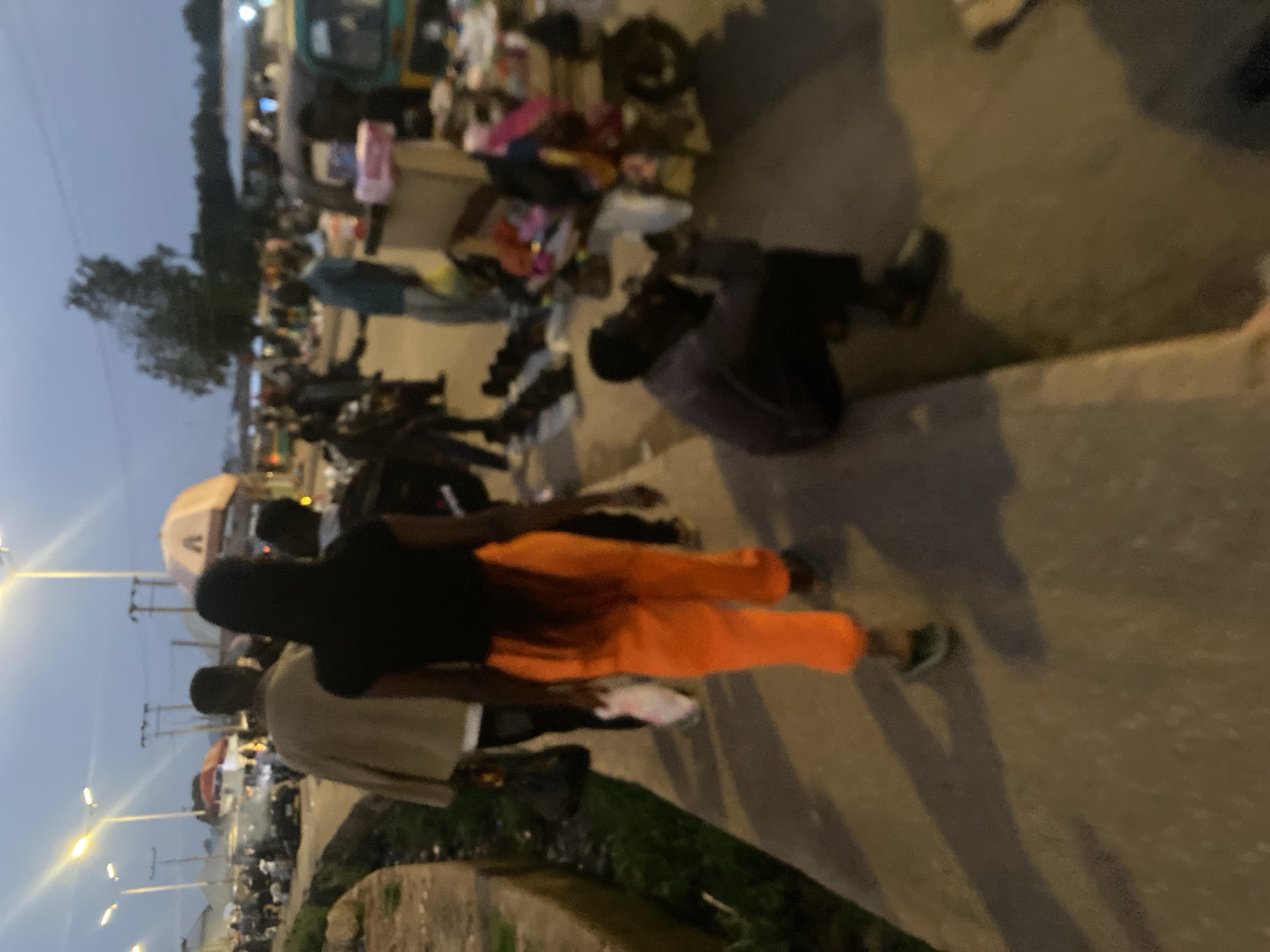 This is a photograph taken in the city of Bukuru, Nigeria, capturing a scene at dusk with limited visibility. The image depicts a street or market area where several people are present, engaging in various activities.
In the foreground, a person is walking away from the camera, wearing bright orange pants and a black top. To their left, another person in a light-colored jacket is also walking, carrying a bag. Further down the path, a person is seated and appears to be talking on a phone or interacting with an object in their hands.
The environment suggests a public space with several stalls or vendor setups. Various objects, possibly goods for sale, are visible on tables and on the ground, including what appear to be shoes and various retail items. Streetlights are illuminated, casting a dim glow over the scene and creating elongated shadows on the paved surface. In the background, more people and vehicles are discernible, hinting at a bustling atmosphere. The sky is a dusky blue, indicating it is either early evening or late morning. The overall impression is that of a busy marketplace at the close of day.