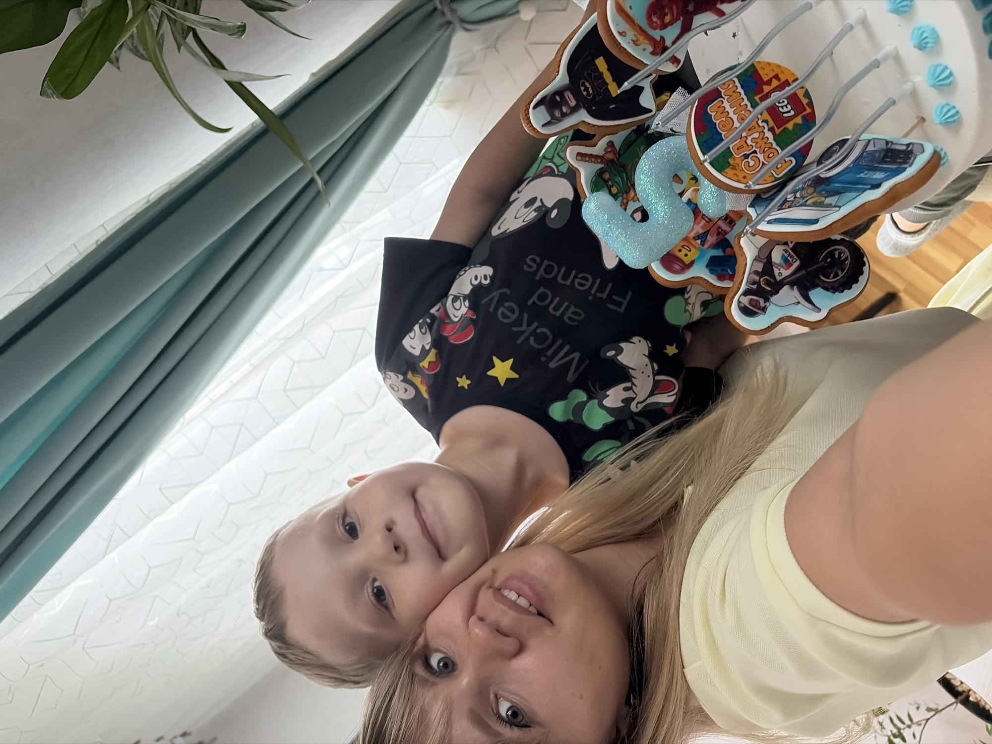 This is a close-up, low-angle selfie of a woman and a young boy, likely mother and son, nestled together. The image is oriented vertically, with the subjects' heads near the bottom. The woman, with blonde hair and light-colored eyes, is looking directly at the camera with a slight smile. The boy, also with blonde hair and blue eyes, is positioned behind and slightly to the left of the woman, also looking towards the camera. He has a gentle smile.
They are in an indoor setting, with a white, textured curtain providing a backdrop. To the upper left, a plant with long green leaves is visible. In the background, there is a white tiered cake decorated with a variety of themed cookies. The cookies appear to be decorated with characters and objects, including what looks like Batman, LEGO figures, a race car, and a motorcycle. There is also a sparkly blue number "2" on the cake. The overall mood is warm and affectionate.
There is visible text on the boy's black t-shirt. It reads "Mickey and Friends" in a playful font, along with images of Mickey Mouse and other cartoon characters. Another cookie on the cake also has visible text in Cyrillic script, which appears to be related to LEGO. The lighting suggests it is daytime, likely indoors with natural light. The scene feels personal and celebratory, possibly for a birthday.