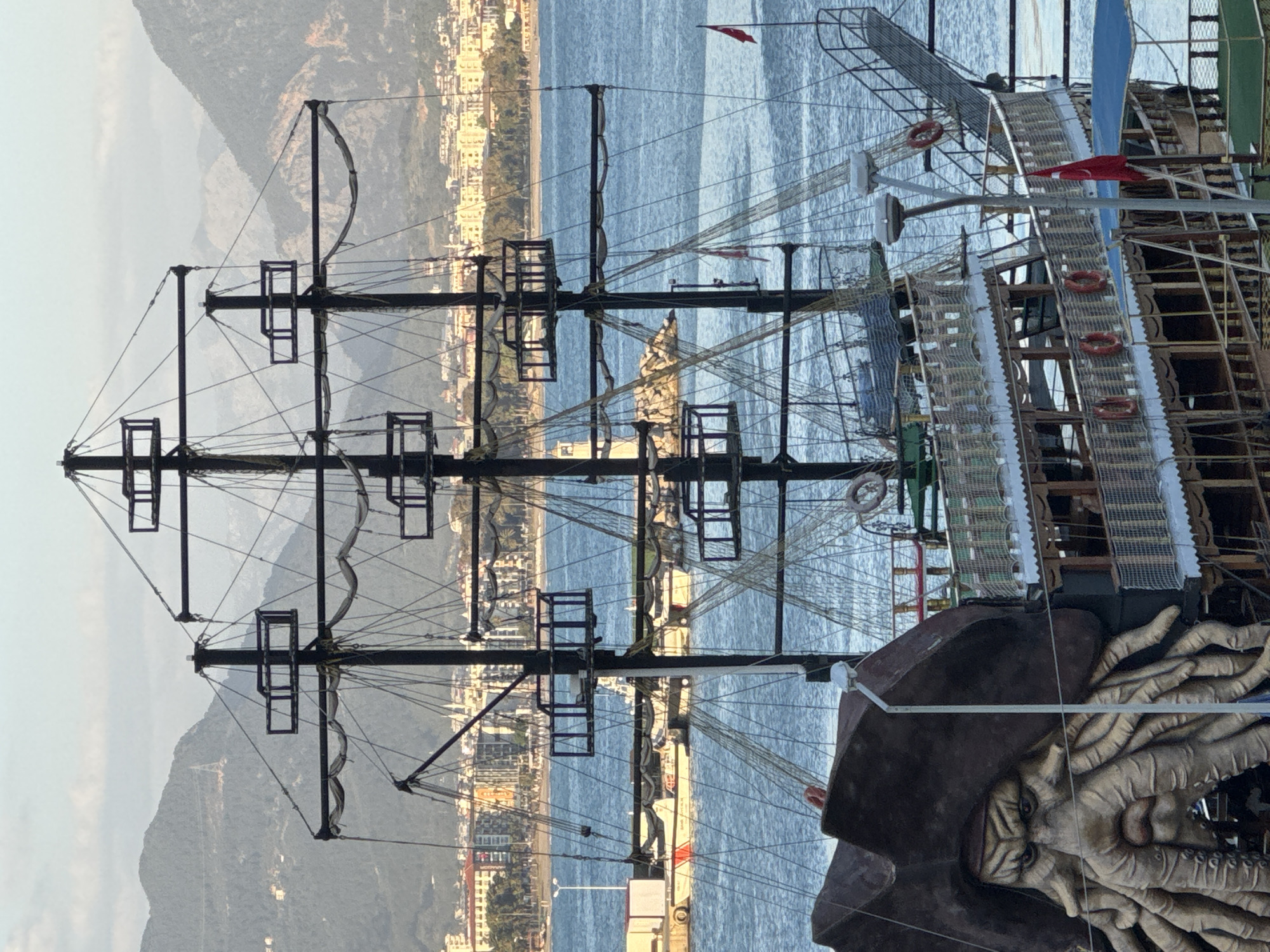 The image depicts a portion of a pirate-themed ship in Alanya, Turkey, with a detailed mast and rigging dominating the foreground. A figurehead resembling Davy Jones from "Pirates of the Caribbean," complete with octopus tentacles, is visible. The sea stretches into the background, with the cityscape of Alanya visible along the shoreline. Mountains form a backdrop to the city. The Turkish flag can be seen flying on the ship. Lifebuoys are attached to the ship's railings.