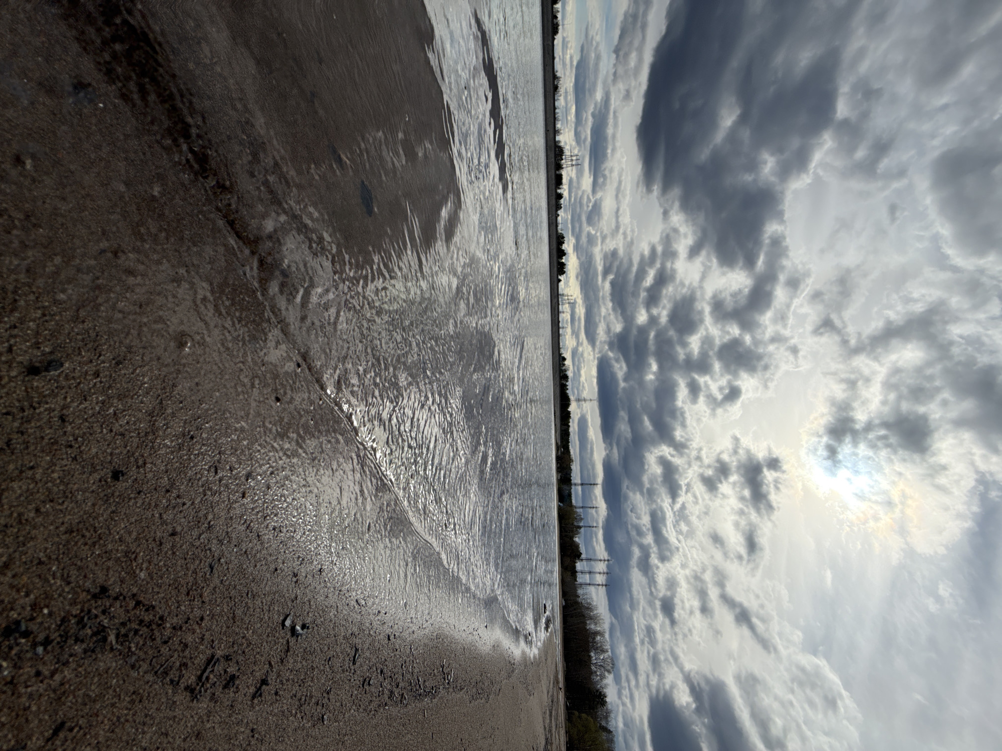 The image shows a shoreline with waves lapping onto the sand. In the distance, power lines are visible. The sky is filled with dark, dramatic clouds with a bright sun partially obscured behind them. The image is oriented sideways. The location is the city of Chaikovsky, Russia.
