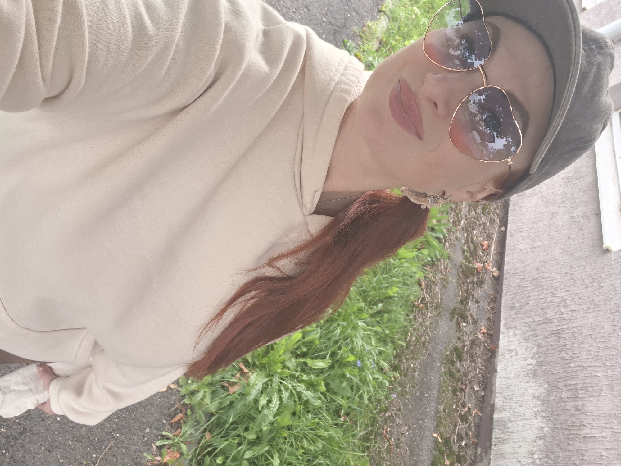 A woman with auburn hair pulled into a ponytail, wearing a light-colored hoodie and a dark baseball cap, poses for a selfie outdoors. She sports heart-shaped sunglasses with rose-gold frames and subtle makeup, including lipstick. A silver-toned earring with decorative elements is visible on her right ear. The background suggests an urban or suburban setting, with a textured wall of a building and a grassy area with some fallen leaves and patches of grey asphalt. The overall impression is of a casual, everyday moment captured in Myadzyel, Belarus.