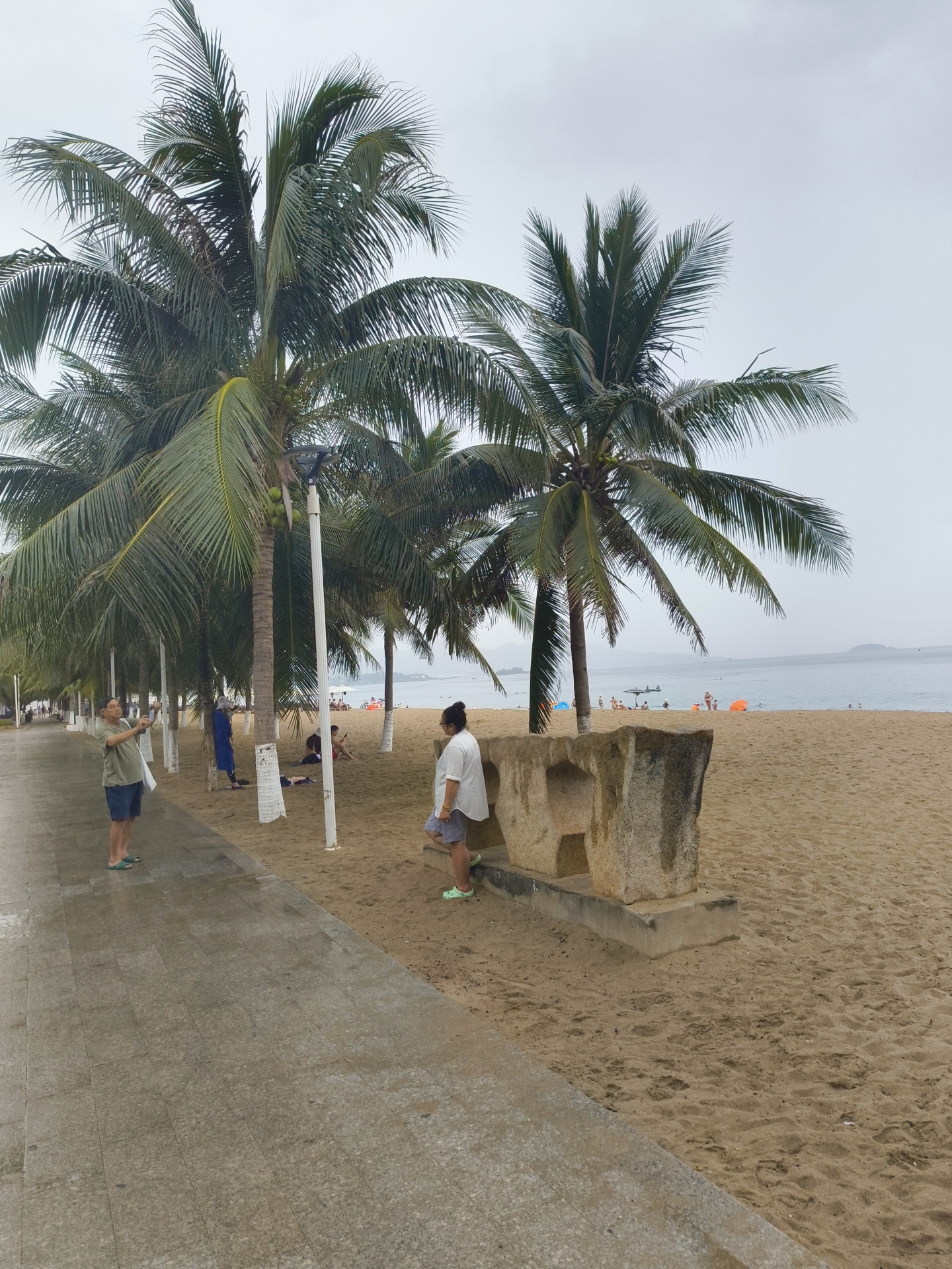 The image depicts a beach scene in Nha Trang, Vietnam, on an overcast day. A paved walkway runs along the left side of the image, wet from recent rain. Several palm trees line the edge of the sandy beach. In the foreground, two individuals are visible. On the left, a man in a green t-shirt and blue shorts stands on the walkway, holding a phone as if taking a photo. To his right, a woman in a white t-shirt and patterned shorts stands on the sand, facing away from the camera. Behind them, several other people are scattered on the beach, some lying down, and others sitting. In the background, the ocean stretches out to the horizon, with a few distant islands or landmasses visible. Small boats and beach umbrellas can be seen on the water and the sand, suggesting a typical beach day despite the cloudy weather. A large, rough-hewn stone structure with carved-out sections is situated on the sand near the two foreground individuals.