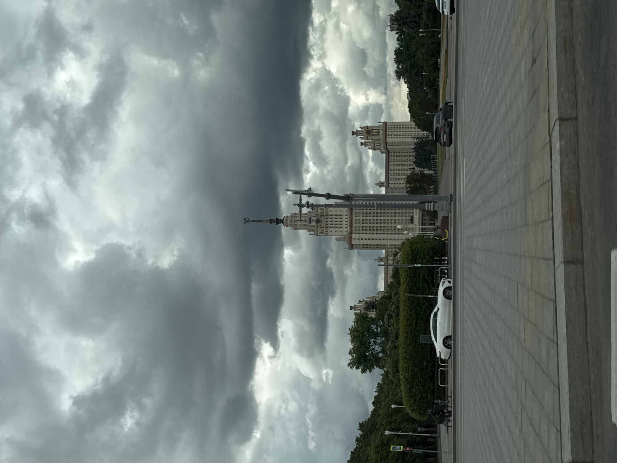 The image shows the main building of Moscow State University (MGU) in Moscow, Russia. The building is a Stalinist skyscraper, with a prominent central tower and flanking wings. The sky is overcast with dark, heavy clouds. A wide paved area leads up to the building, with trees lining the edge. Cars are parked along the side of the road.