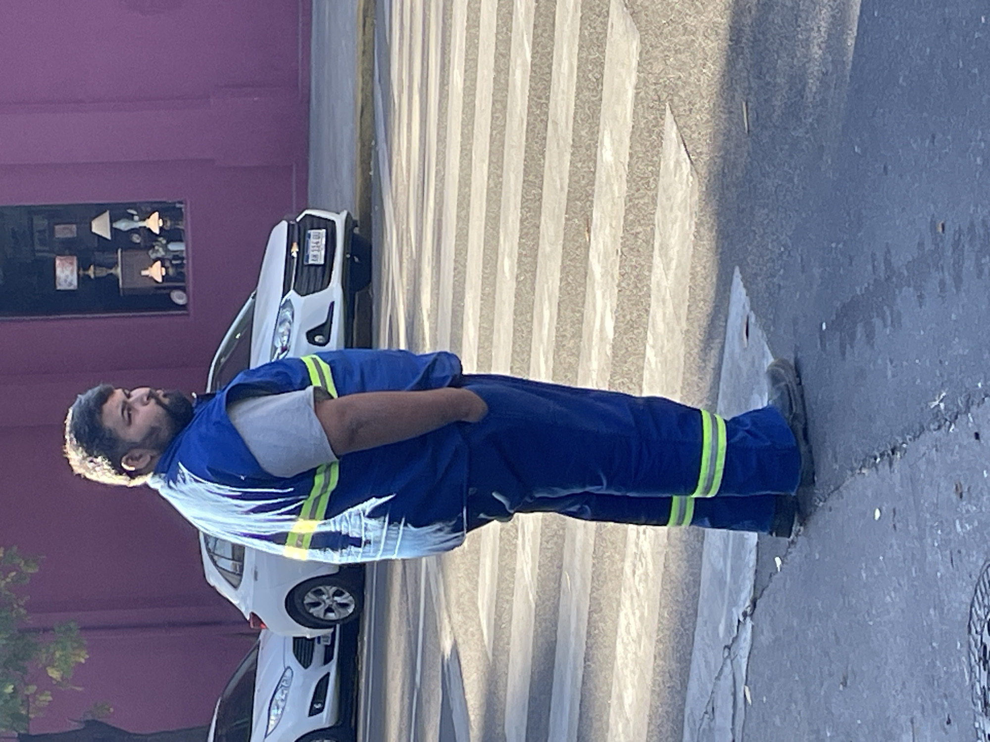 An adult man stands on a sidewalk next to a pedestrian crossing in Buenos Aires, Argentina. He is captured from the waist up, facing towards the right side of the frame with his head turned slightly upward. He has dark hair and a beard, with a subtle highlight on the left side of his hair, suggesting strong, low-angle sunlight. The man is wearing a blue uniform with bright yellow-green reflective stripes on the shoulders, waist, and lower legs, over a grey t-shirt. His hands are in his pockets.
Behind the man, a white car, possibly a Chevrolet Onix or similar model, is parked facing left. Its license plate is partially visible, displaying "AB 134 QO". Another car is partially visible behind the white one. To the left of the man, a striking purple building wall is visible, featuring a window that reflects several ornate lamps or chandeliers. The ground features a textured pavement and a clear zebra crossing (pedestrian crossing). Shadows are long and distinct, indicating a sunny day, likely during the morning or late afternoon. A metal manhole cover is partially visible on the right side of the road. The overall scene depicts a typical urban street environment.