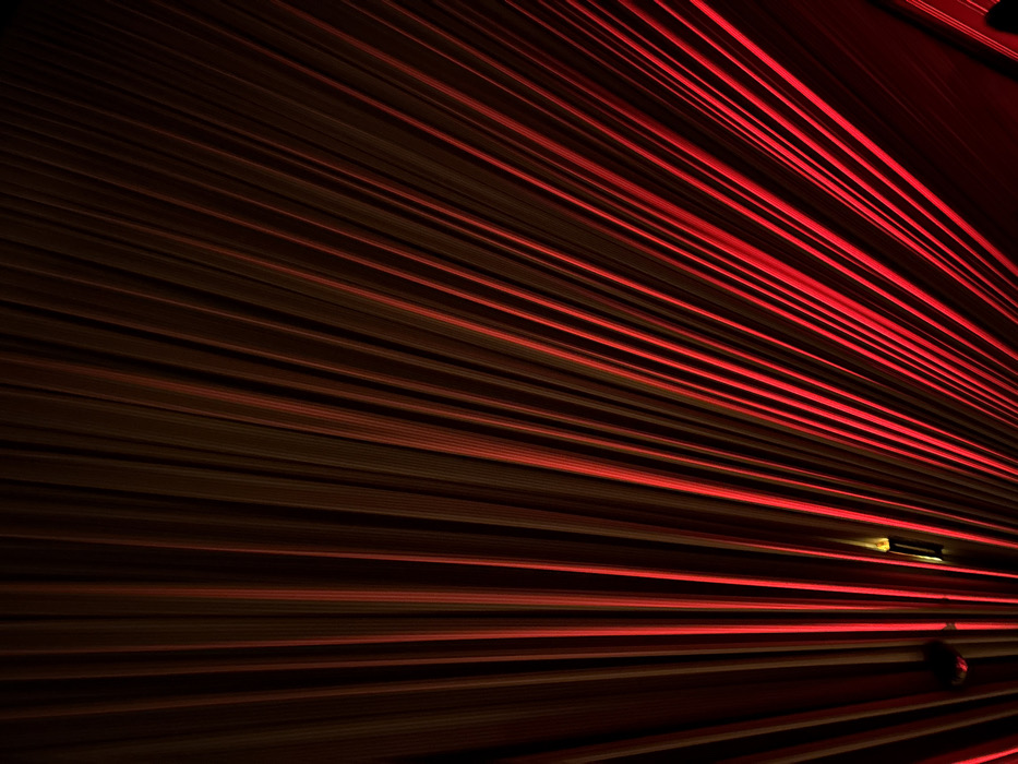 The image presents a close-up view of a surface composed of numerous parallel lines. These lines alternate between dark brown and bright red, creating a striated pattern that converges towards the lower right corner. The lighting emphasizes the linearity of the texture, casting long shadows and highlighting the raised edges of the lines. A small, dark object is visible near the center-right of the frame. The overall effect is abstract and dramatic, using color and perspective to create a sense of depth and movement.