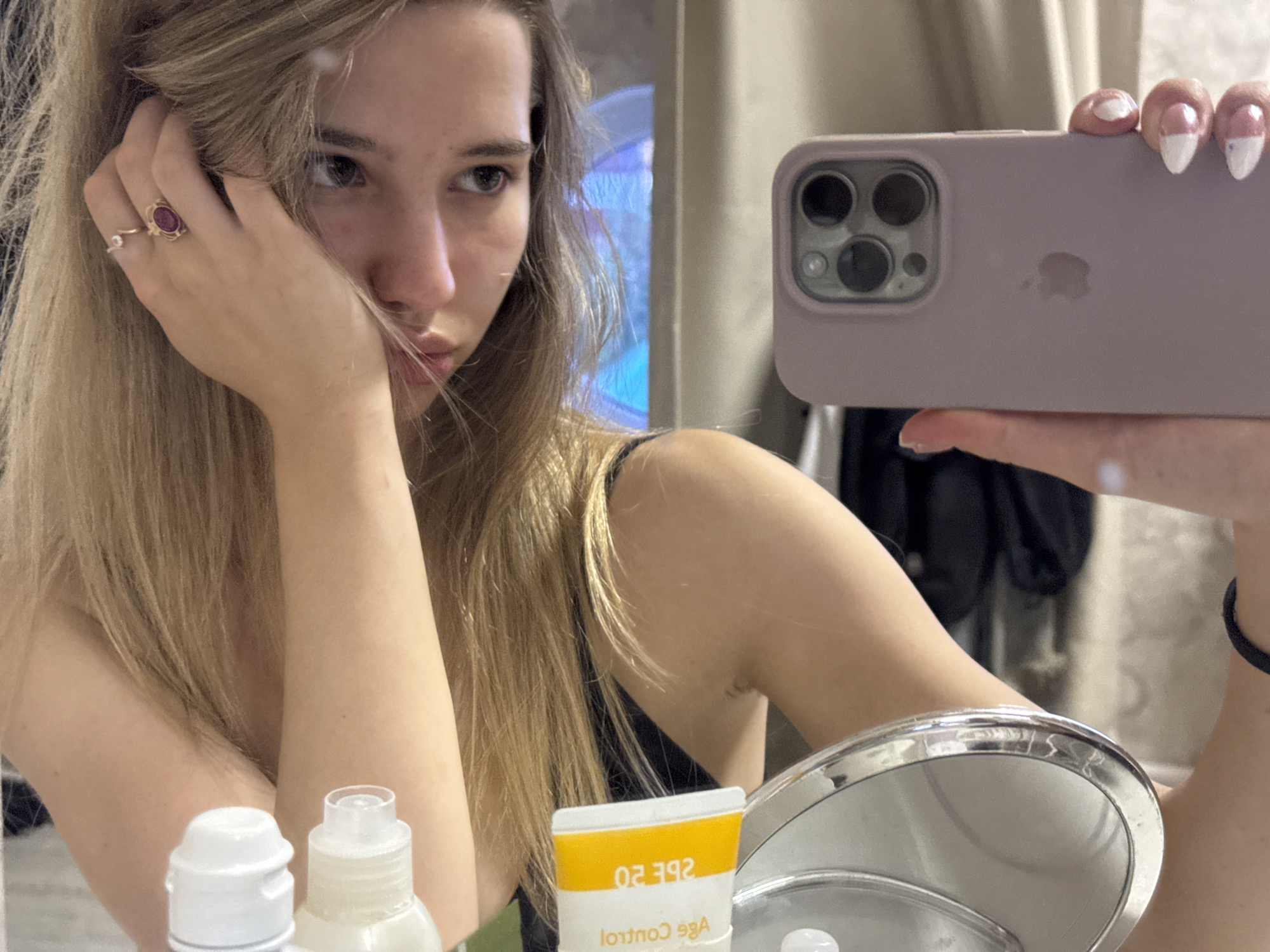 A young woman is captured in a mirror selfie, showing her reflection from the chest up. She has long, light brown hair that falls around her face and shoulders. Her left hand is resting against her cheek, with hair partially covering her face. Two rings are visible on her left ring finger: one with a prominent oval-shaped red or purple gemstone, and a simpler band. She appears to be wearing a dark, possibly black, top or camisole.
Her gaze is directed to her right, away from the camera's lens, giving her a pensive or contemplative expression. Her right hand, with nails painted with white tips, holds a light purple iPhone in a matching case. The phone's triple camera array and the Apple logo are clearly visible in the reflection. A black scrunchie is worn on her right wrist.
The setting appears to be an indoor space, possibly a bathroom or dressing area, indicated by cosmetic products in the foreground. On the left, two white bottles with white caps are partially visible. To the right, a yellow and white tube reflects text that, when un-mirrored, reads "SPF 20" and "Age Control". A smaller, round vanity mirror with a chrome rim is situated in the bottom right corner. The background reflection shows what looks like a light-colored wall or curtain, with a faint blue light source visible behind the woman.