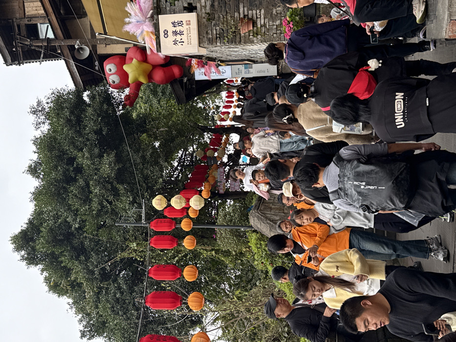 The image depicts a crowded outdoor pedestrian area within Yuzhong District, China, under an overcast sky. The scene shows a sloping pathway flanked by traditional-style architecture on the right and dense foliage on the left.
Numerous individuals, primarily East Asian adults with some children, occupy the pathway, moving in various directions, though a general upward flow is evident. Most are dressed in casual attire. In the foreground, a man in a black long-sleeved shirt is visible on the left. To his right, a woman in a yellow cardigan holds an object. Another woman in an orange jacket stands in the mid-left, focused on a mobile phone. A man wearing a grey t-shirt and a black backpack with white branding is prominent in the center foreground, facing away from the viewer. Further back, individuals are seen walking and standing, with some also engaging with mobile devices.
Overhead, multiple strings of decorative lanterns are suspended, featuring both cylindrical red lanterns and spherical orange-yellow lanterns. These lanterns extend into the background. A large, dense green tree canopy dominates the upper left and central background.
On the right side of the pathway, a multi-story building with traditional Chinese architectural elements is visible, featuring dark wooden structures, a tiled roof, and a stone wall base. A large red inflatable cartoon character with round eyes and a yellow star in its hand is affixed to the upper facade of this building. Below the inflatable, a white sign with Chinese characters "外婆桥 小食店" and "WAIPOQIAO SNACK SHOP" in English is mounted on the stone wall. A utility pole with wires and a loudspeaker is also attached to the building. Potted plants are visible on ledges along the stone wall. The ground appears to be a paved surface, likely stone or concrete.