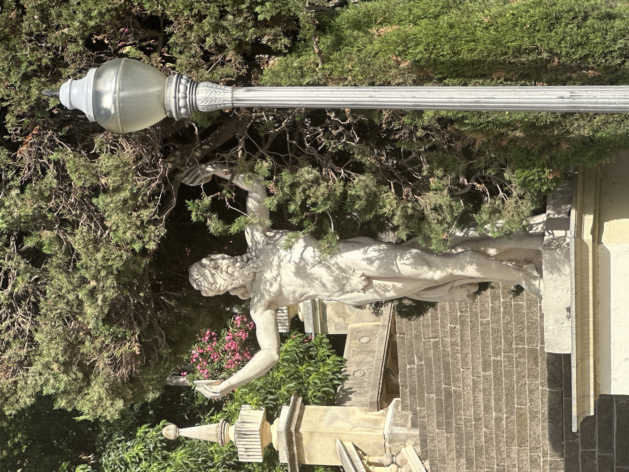 The photograph depicts a sun-drenched outdoor scene featuring a classical stone statue of a male figure with a beard, standing on a pedestal. The statue is partially obscured by lush green foliage and a tall, ornate lamppost with a silver finish. Behind the statue and to the left, a stone staircase with multiple steps leads upwards, flanked by a stone balustrade and a decorative finial. Pink flowers are visible in the background, adding a splash of color to the scene. The lighting suggests it is daytime, with strong shadows cast by the trees and the lamppost.
This scene is located in the Parc de la Ciutadella in Barcelona, Spain, specifically near the Cascada Monumental fountain, where numerous statues and decorative elements are present. The statue is likely a mythological figure, consistent with the decorative style of the park. The lamppost is a typical fixture for park pathways. The environment is an urban park setting, with well-maintained landscaping and architectural features.