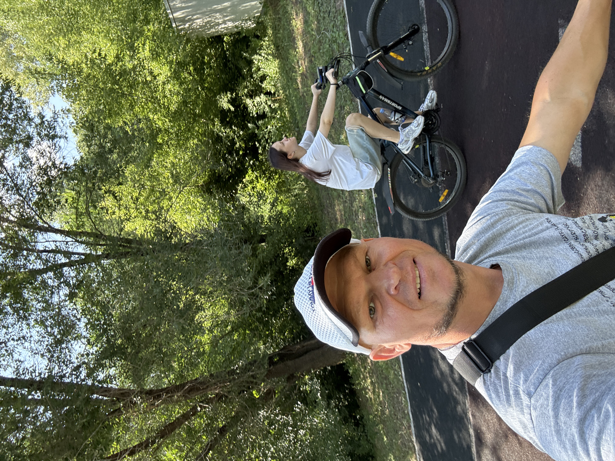 In the city of Oskemen, Kazakhstan, a man in a grey t-shirt and a white baseball cap takes a selfie on a sunny day, with a lush green forest in the background. To his right, a young woman with long brown hair rides a bicycle on a paved path. She wears a white t-shirt and light-colored shorts, her gaze directed upwards and to the right. The bicycle is dark blue and has the brand name "SPORTSMAN" visible on the frame. A water bottle is attached to the frame. The ground is dark asphalt, suggesting a park or recreational area. The overall atmosphere is one of outdoor activity and leisure.