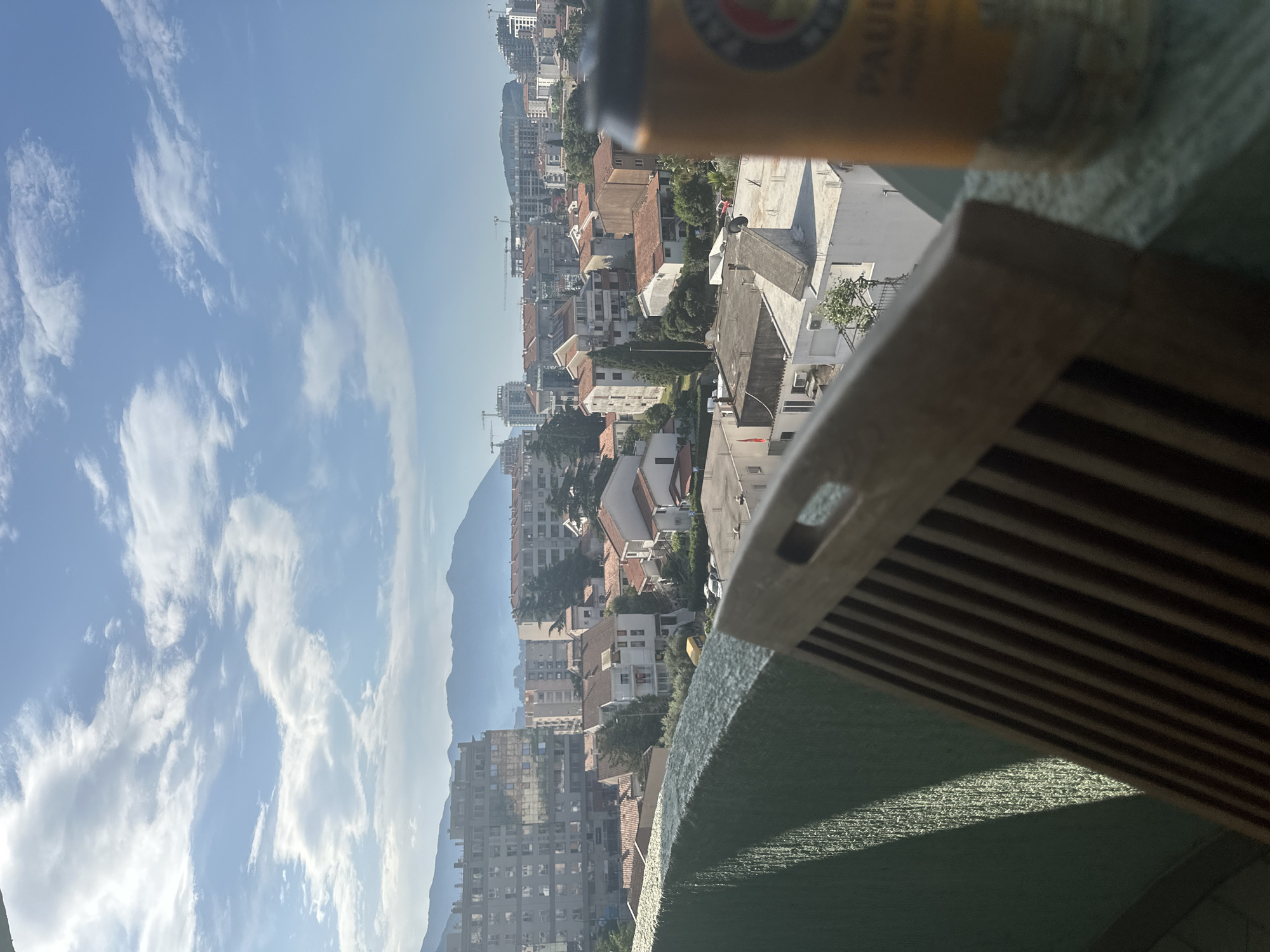 The image shows an elevated view of the city of Budva, Montenegro, from a balcony. In the foreground, there is a partial view of a wooden structure and a beer can. The background reveals a dense cityscape with residential buildings, trees, and a mountain range in the distance. The sky is partially cloudy, indicating a bright, sunny day. Several construction cranes are visible, indicating ongoing development.