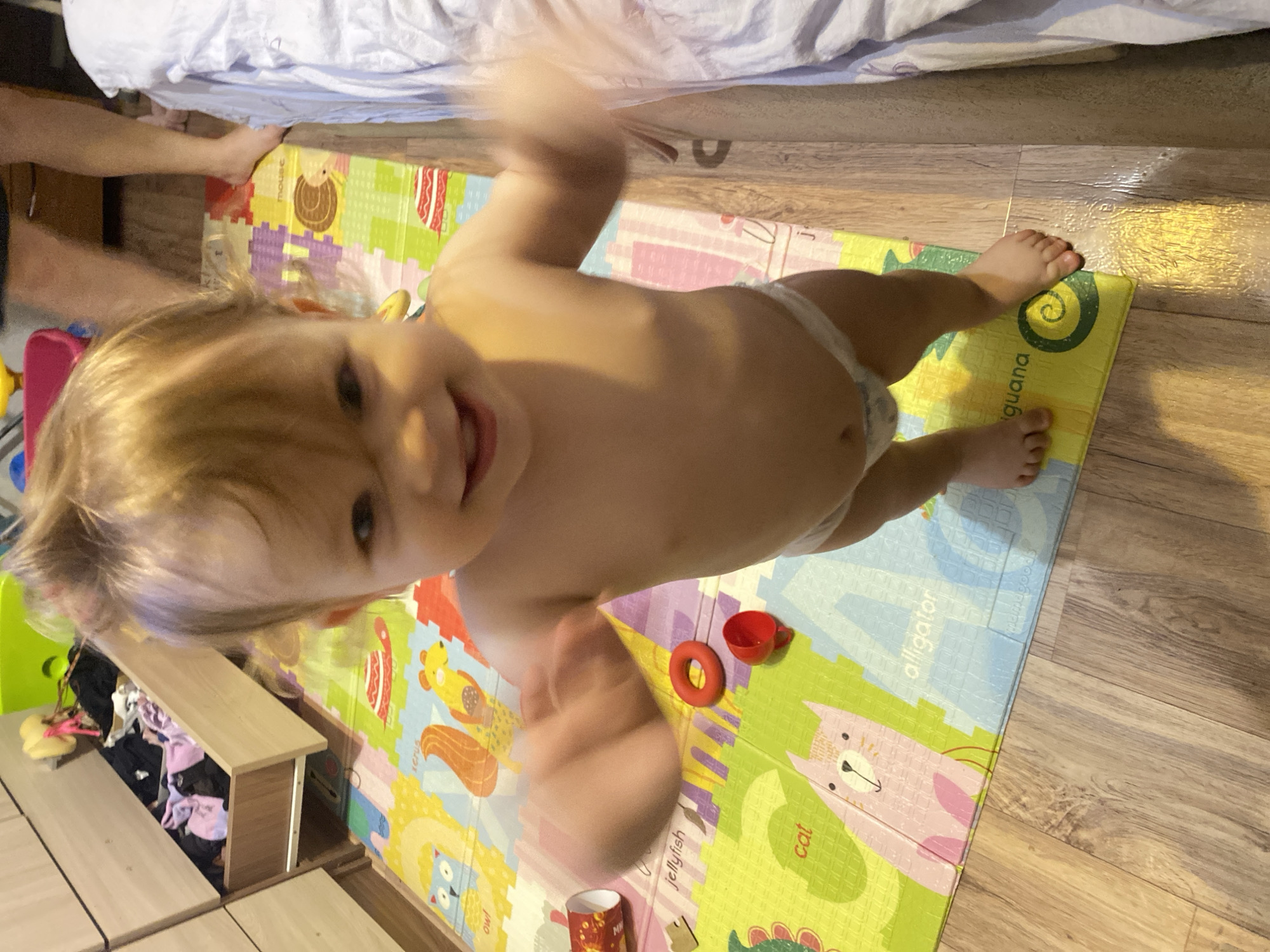 A close-up, high-angle, tilted indoor shot shows a toddler waving in a blurred motion with a happy expression, standing on a colorful alphabet playmat with toys. The mat features images of animals and objects labeled with words like "iguana," "cat," and "alligator." To the side of the mat is a wooden floor. A bed and a dresser are partially visible, along with a glimpse of an adult's feet near the edge of the playmat. The location appears to be a child's playroom or bedroom. The city is Unknown, Unknown.