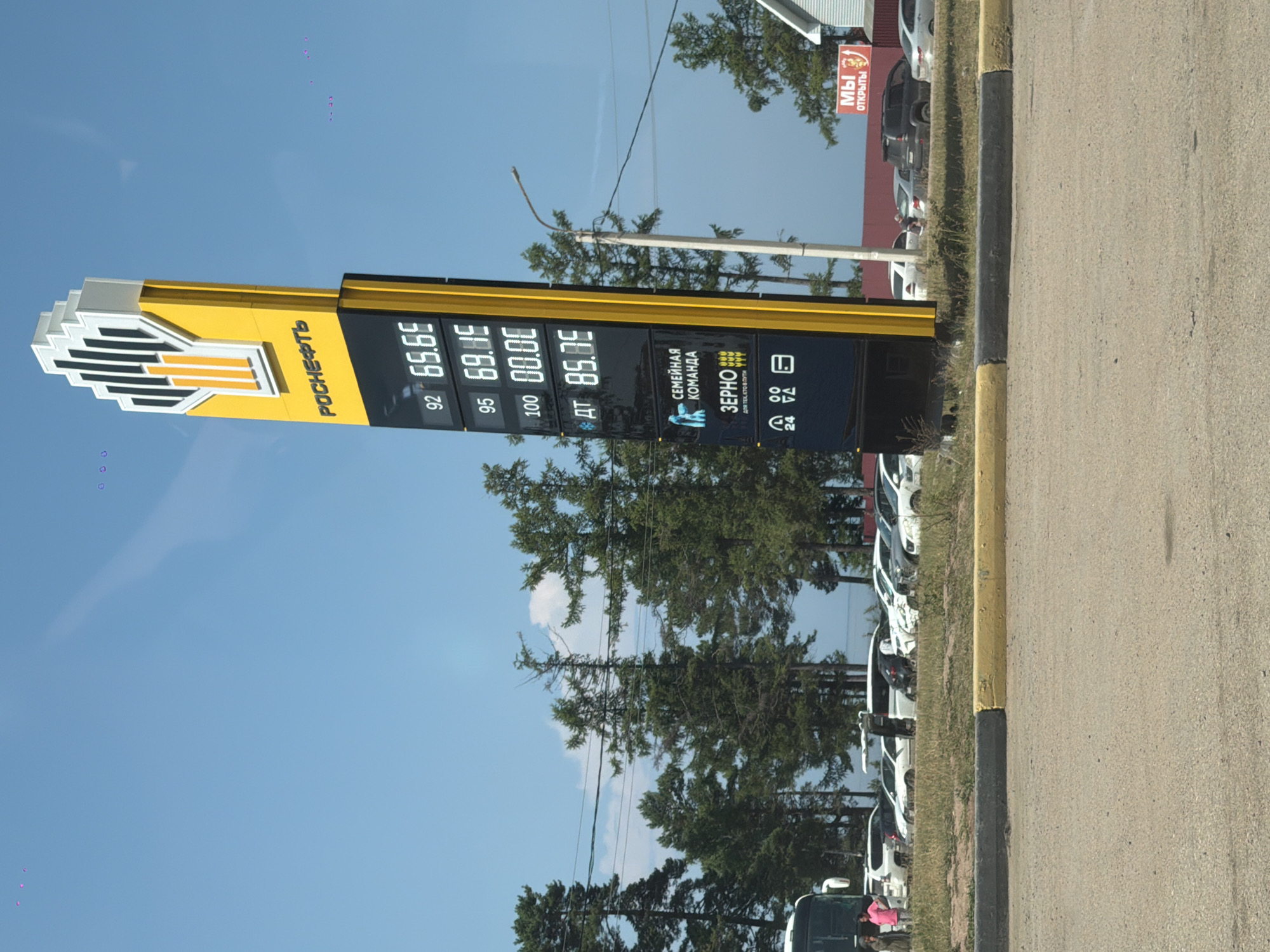 A tall, bright yellow and black signpost displays fuel prices at a Rosneft gas station under a clear blue sky. The sign shows prices for 92 octane fuel at 65.65, 95 octane at 69.15, 100 octane at 00.00, and diesel (AT) at 85.95. Below the fuel prices, there are logos for "SEMEINAYA KOMANDA" (Family Team) and "ZERNO" (Grain), along with icons indicating 24-hour service and other amenities.
In the background, a line of parked cars, including SUVs and sedans, are visible, as well as a white bus. A few people are seen standing near the bus on the left side of the image. The gas station is situated near a row of coniferous trees, and a street light stands to the right of the price sign. A red sign with white lettering that reads "МЫ ОТКРЫТЫ" (We are open) is visible on a building in the far right background. The foreground consists of a paved area and a concrete curb painted yellow and black. The overall scene suggests a roadside service area in Turka, Russia.