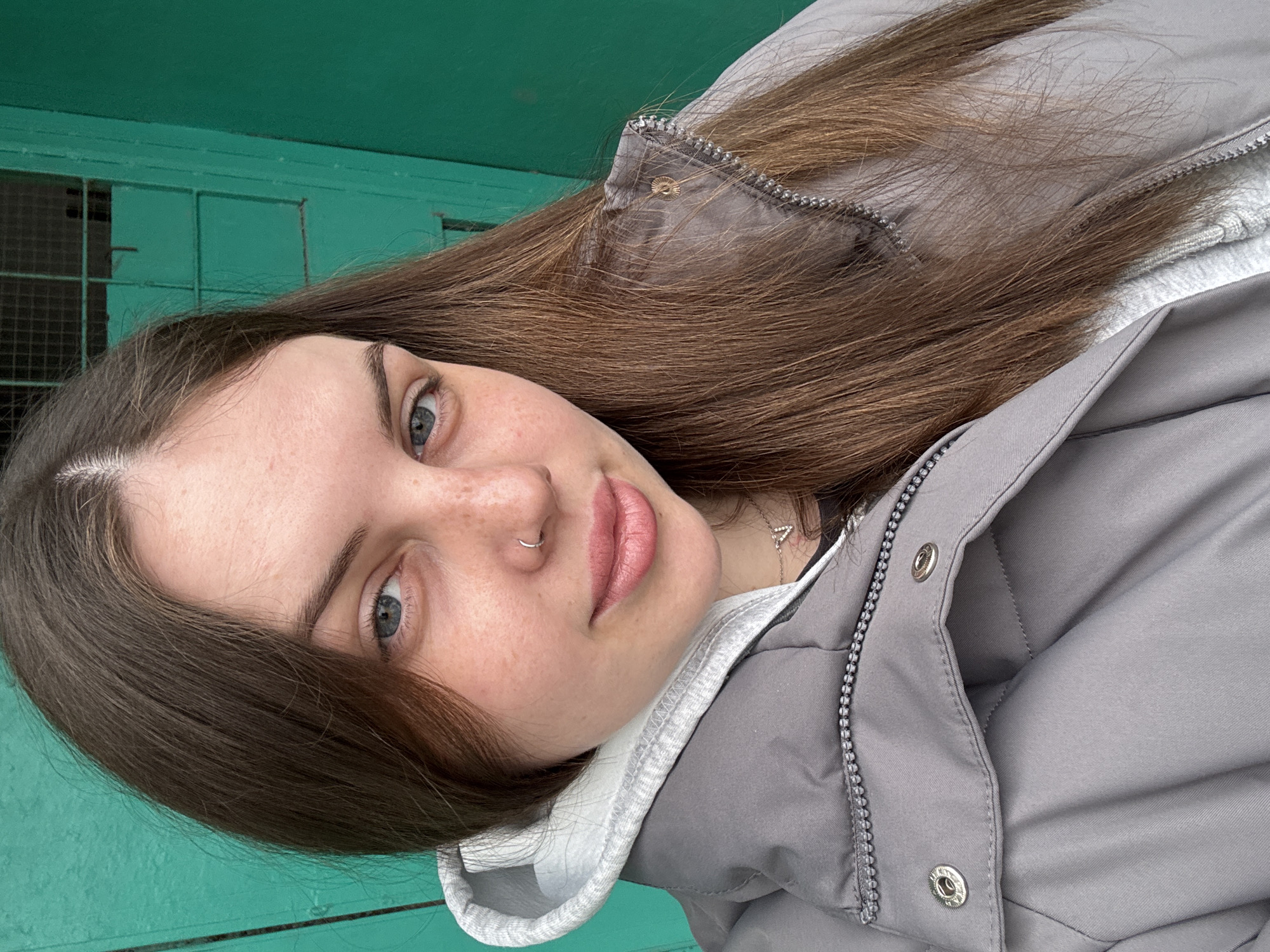 A young woman with long, brown hair and light eyes is looking at the camera. She has freckles on her face and is wearing a nose ring. She is wearing a gray jacket with a white hoodie underneath. The background appears to be a green painted structure, possibly a building or fence. The location may be Chornomorskoe, Ukraine.