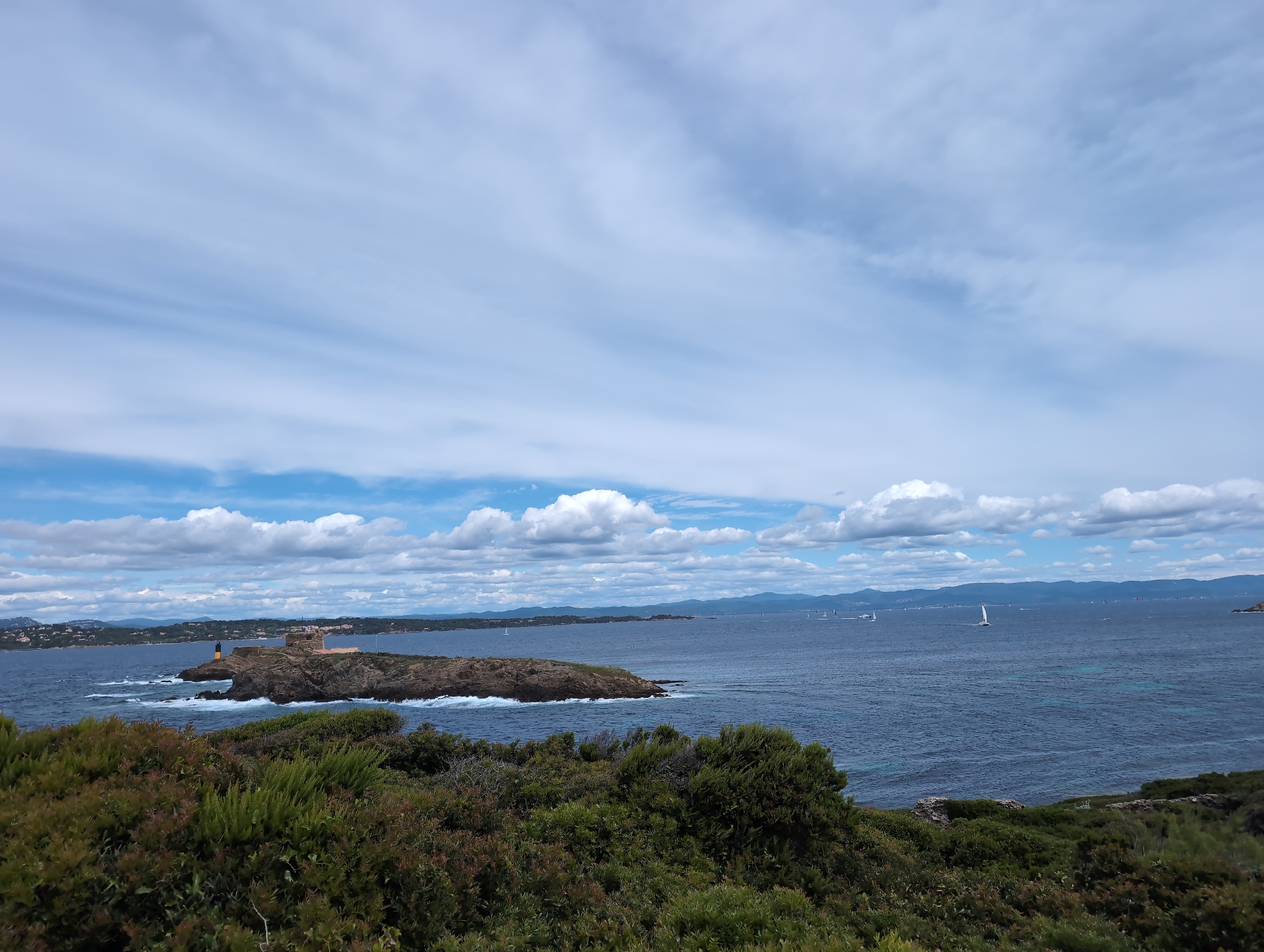 A landscape photograph captures a coastal scene, possibly taken near Hyères, France, featuring a small, rocky island with a building on it in the middle distance, viewed from a shrub-covered foreground. The island exhibits a light-colored structure with multiple sections, possibly a historic tower or fort. The sea appears to be a deep blue, with a few small white sailing vessels visible in the distance. The sky is partly cloudy, with layered white and grey clouds partially obscuring the blue. The vegetation in the foreground is lush and green, with dense bushes covering the land. There is no visible human presence in the photo other than the structure on the island.