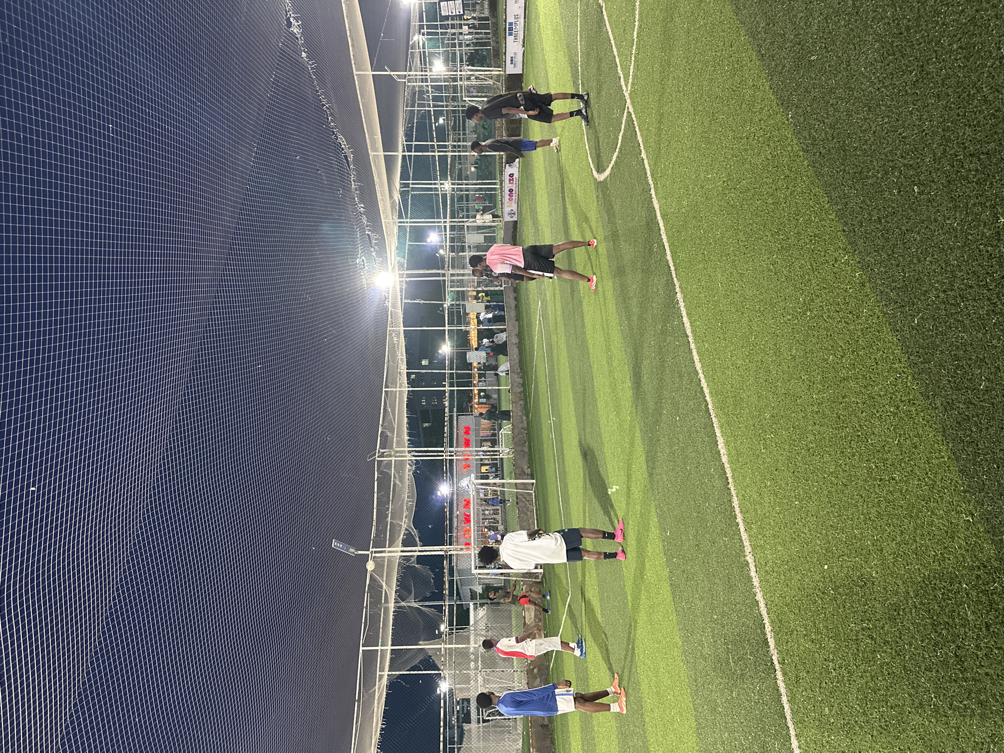 An artificial turf soccer field is illuminated by stadium lights under a dark sky with a net overhead, likely in Abuja, Nigeria. Several men are scattered across the field, playing a game. In the foreground, on the left, a man in a blue long-sleeved shirt and white shorts with orange accents stands facing away from the viewer. To his right, another man wears a white shirt with red accents on the sleeves and collar, and white shorts. Further back, a man in a white t-shirt and black shorts is visible. On the right side of the frame, a man in a pink t-shirt and black shorts faces away, and to his right, a man in a black t-shirt and black shorts is also present. In the background, a goal is set up, and other figures are visible near the sidelines and in the distance, suggesting spectators or other players. Banners are affixed to the fencing around the field, including one with the text "MONOIZA CYBER FORENSICS" and another with "THREEMPLUS."