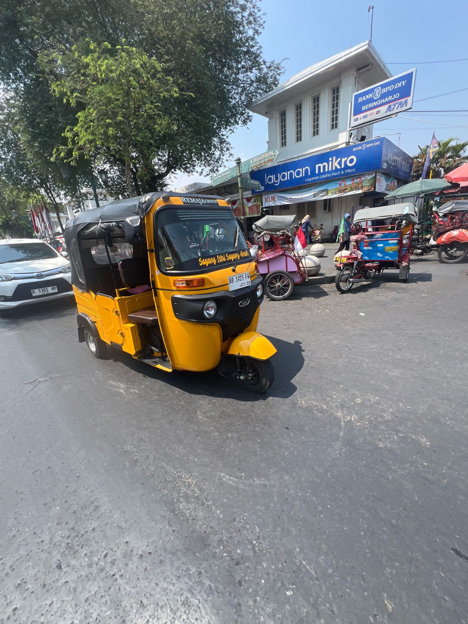 The image depicts a street scene in Yogyakarta, Indonesia, likely in an urban area given the presence of businesses and motorized vehicles. In the foreground, a yellow three-wheeled auto-rickshaw, known as an "ojek" or "becak motor" in Indonesia, is prominently featured. The vehicle has a black canopy and visible seating. Behind it, a white car with a license plate visible, "R 1381 NP," is parked.
In the background, a multi-story building stands, with signs indicating it houses "BANK BPD DIY" and "layanan mikro" (micro services). These businesses suggest a commercial district. Several other similar auto-rickshaws are also present, some appearing to be in use or parked. People are seen walking in the area, and the overall environment suggests a bright, sunny day with clear skies. The road surface appears to be asphalt, showing some wear and tear. Trees line the side of the street, contributing to the urban but somewhat tropical feel of the location. The scene captures a typical snapshot of daily life and transportation in a bustling Indonesian city.