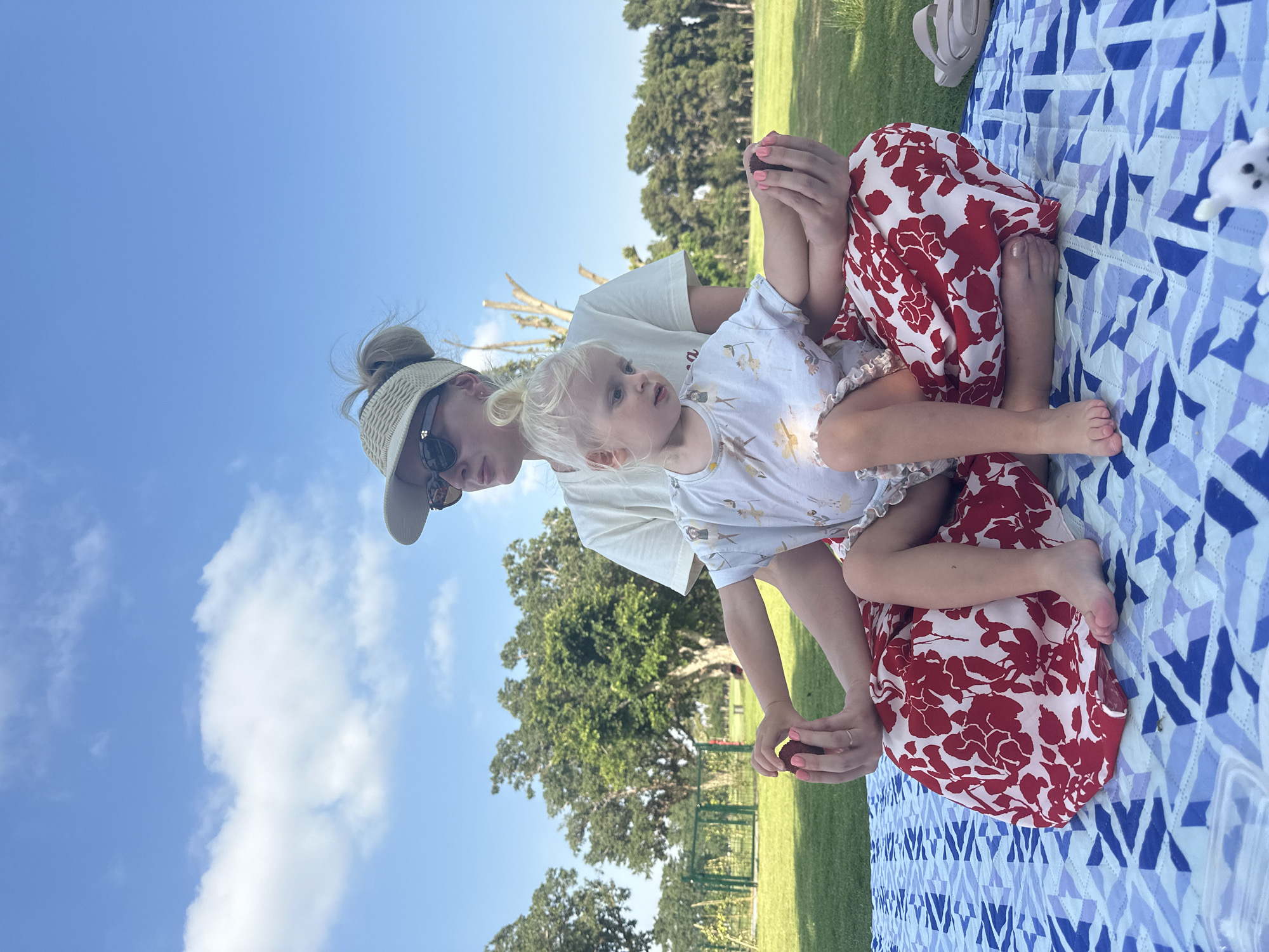 A woman and a young child are sitting on a picnic blanket in a grassy park. The woman is wearing a white visor, sunglasses, and a white t-shirt. The child is wearing a white romper with a subtle pattern and has blonde hair in a bun. The child is sitting on the woman's lap, and both are looking down. The woman is holding a small, dark, spherical object in her left hand, and the child is reaching towards it with their right hand. The picnic blanket has a geometric pattern of blue and white shapes. In the background, there are trees and a clear blue sky with some clouds. The location appears to be a park or a large green space, possibly within the city of Ramat Gan, Israel, given the presence of lush greenery and park-like landscaping.