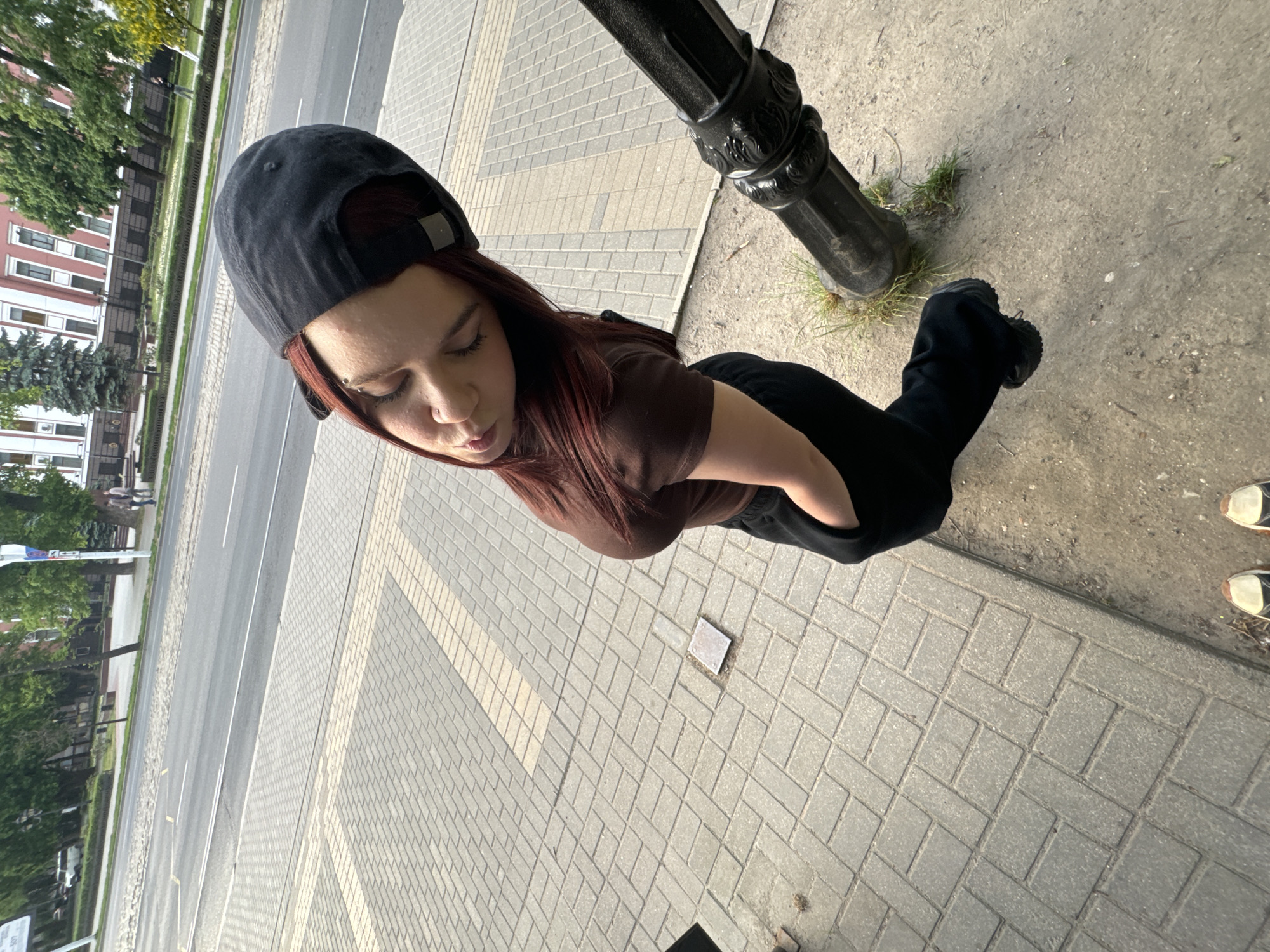 A high-angle shot shows a person in dark clothing and a black baseball cap standing on a brick-paved surface adjacent to a street. The person has dark hair and is wearing a brown top. A black metal lamppost is visible in the upper right of the frame, rooted in the ground at the edge of the pavement. Across the street, there are trees and buildings. The street is paved and has lane markings. Another person's feet are partially visible in the bottom right corner of the image, suggesting the proximity of another individual. The setting is an urban street during daylight.