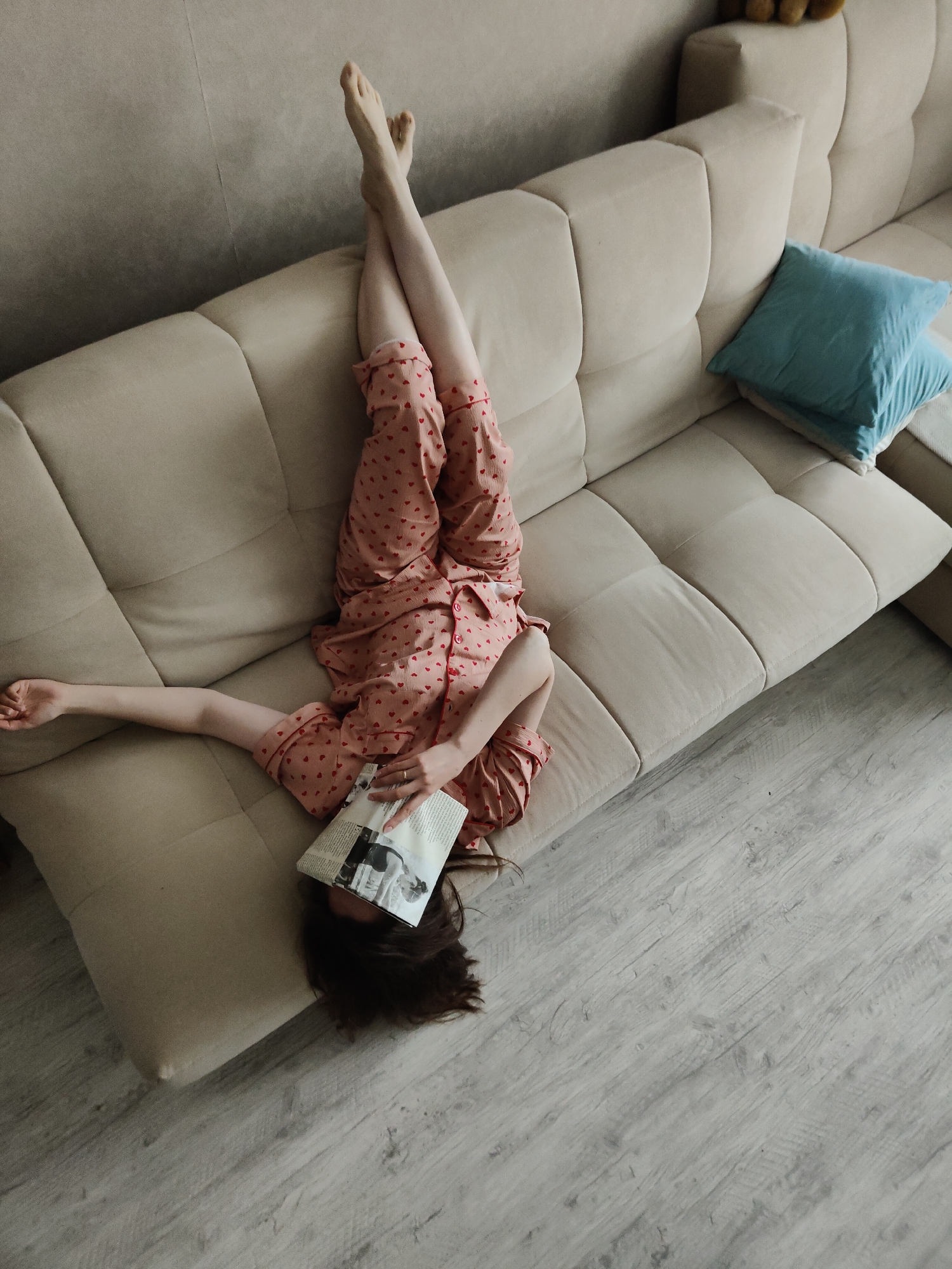An overhead, high-angle shot depicts a person lying upside down on a large, light beige sectional sofa. The person, whose head is positioned at the lower end of the sofa near the floor, has their legs raised and crossed, resting against the backrest of the couch. They are wearing light pink pajamas adorned with small red heart patterns.
An open magazine or book is held over the person's face, partially obscuring it. The pages display some illegible text and a black and white image. A ring is visible on the left hand holding the publication. The person's right arm extends along the sofa cushion.
The sofa is upholstered in a neutral, light beige fabric with segmented seat and back cushions. Two bright blue throw pillows are neatly stacked on the far right section of the sofa. The flooring beneath is composed of light greyish-brown wooden planks, exhibiting a subtle wood grain. A plain, light-colored wall forms the background of the scene. The image evokes a relaxed and leisurely atmosphere, suggesting a moment of casual comfort indoors.