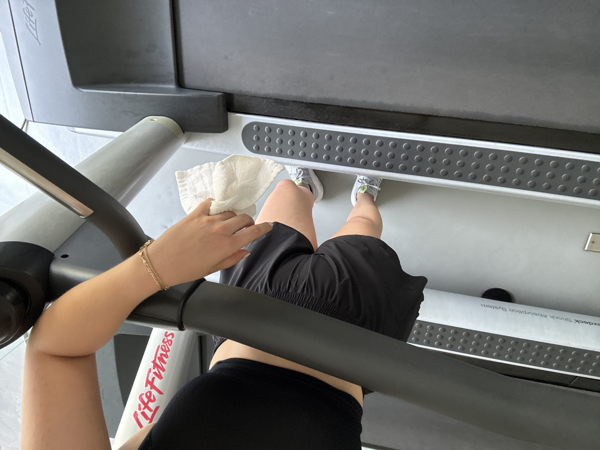 An overhead, close-up shot shows a person standing on a LifeFitness treadmill. The person is wearing black shorts, a black top, and grey sneakers with a touch of green. A white towel is in their left hand, which is also adorned with a gold bracelet. The treadmill features a grey base with a black tread, and the handles are black. The floor is grey. The location is potentially within Mexico City, Mexico.