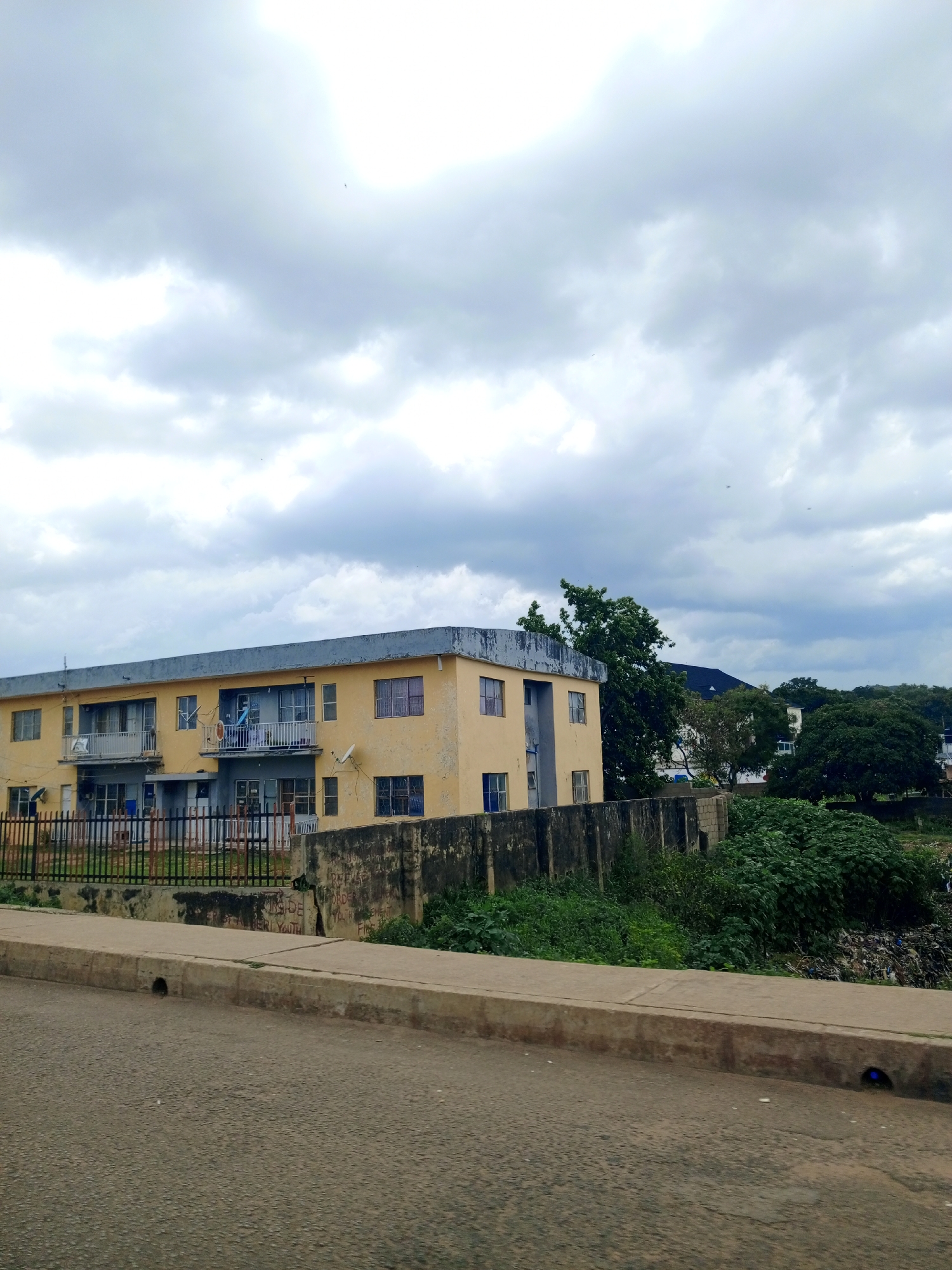 An exterior, eye-level, medium shot captures a weathered, two-story, pale yellow apartment building under a cloudy sky. The building features multiple windows and balconies with railings. A black metal fence stands in front of the building. A concrete wall, some parts graffitied, runs along the property line. Lush green vegetation and trees surround the building and extend into the background. In the far right background, a pile of trash is visible. A paved road with a concrete curb occupies the foreground. The scene is located in Kabong, Nigeria.