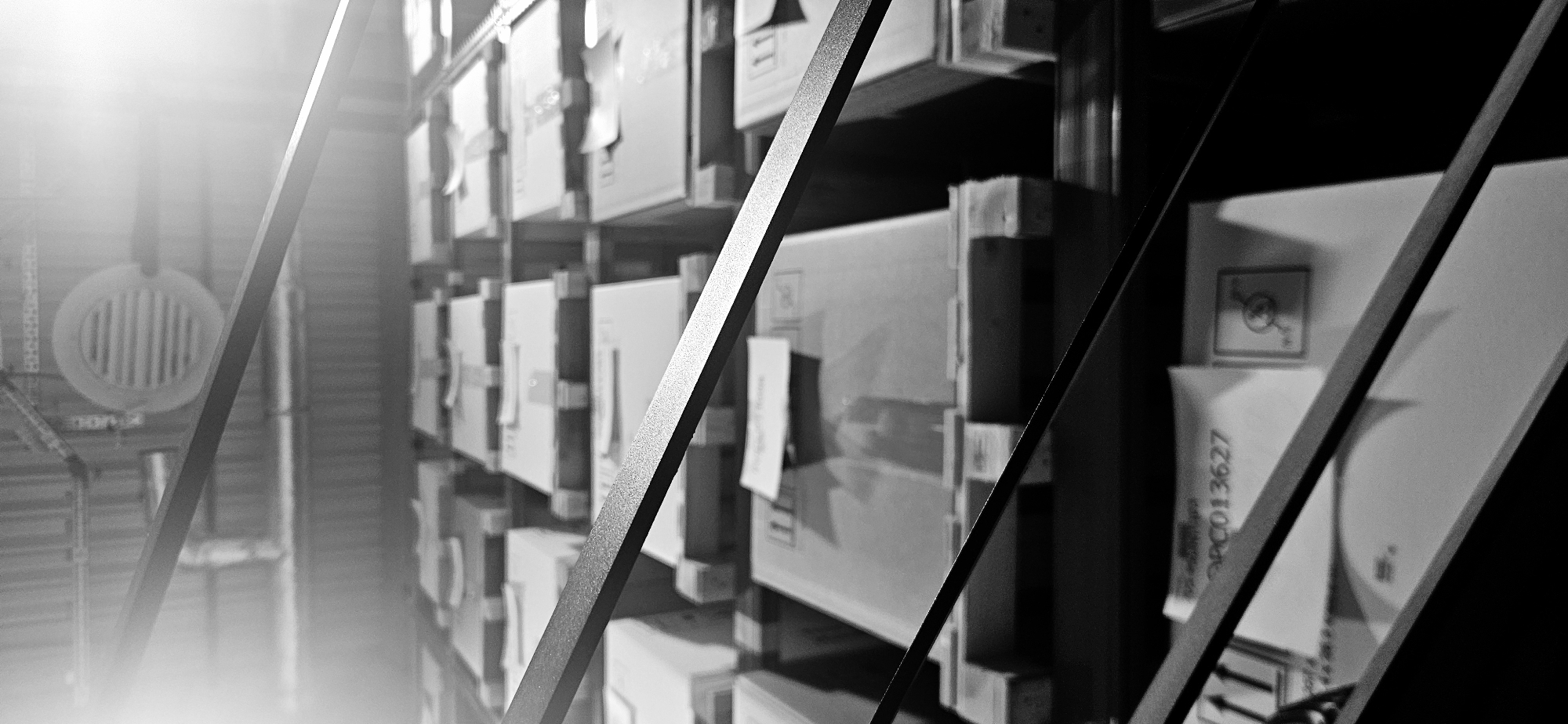 The image shows a storage area, likely an archive or warehouse, containing rows of stacked boxes. The boxes appear uniform in size and shape, suggesting they hold similar contents. Labels or documents are affixed to some of the boxes, presumably for identification and organization. A metal framework, possibly part of the shelving system, is in the foreground. The lighting is subdued, with some areas obscured in shadow. The location is unspecified, so the city of Plovdiv, Bulgaria cannot be confirmed. There are no visible people, actions or interactions present in the image.