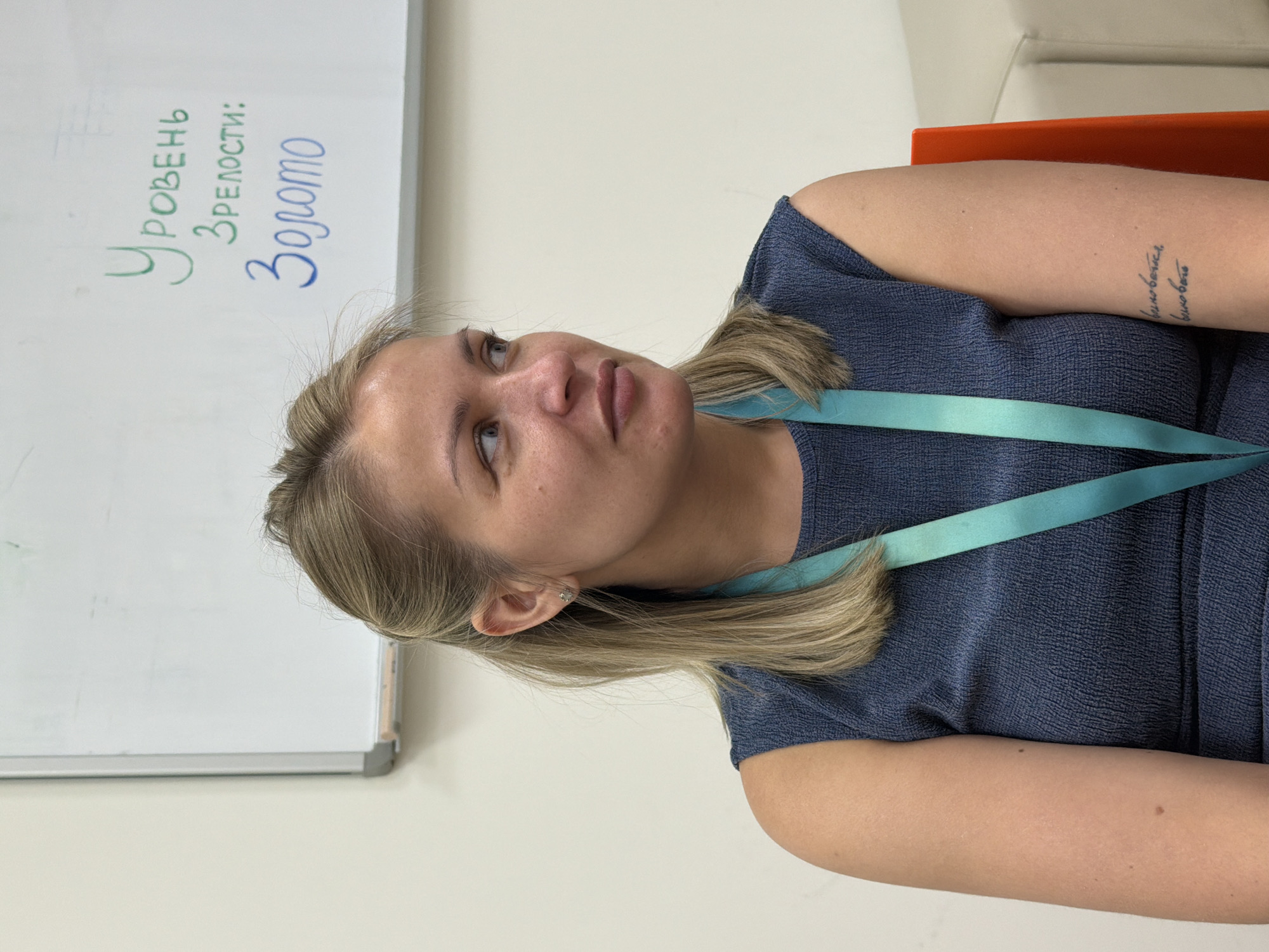 A medium shot captures a fair-skinned woman with blonde hair, a blue dress, and a light blue lanyard. A tattoo is visible on her left arm. Her gaze is directed upwards, towards a whiteboard on the left, which has the words "Уровень зрелости: Золото" written on it in green and blue marker. The background is a plain, light-colored wall. The setting suggests an indoor environment, possibly an office or classroom. There is no explicit information within the image to indicate that this photograph was taken in the city of Krasnoyarsk, Russia.