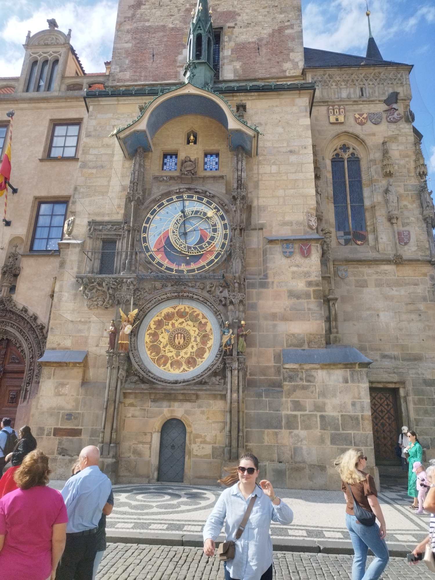 The image shows a bustling street scene in front of the Astronomical Clock in Prague, Czech Republic. The clock tower, a medieval masterpiece, dominates the background, its intricate astronomical dial and figures capturing the attention of onlookers. Several people are visible in the foreground, some pausing to admire the clock, while others are in motion, navigating the cobblestone street. The architecture suggests a historic European city, with stone buildings and a paved plaza. The sky is partly cloudy, indicating fair weather. The scene is lively and suggests a tourist attraction drawing a crowd on a pleasant day.