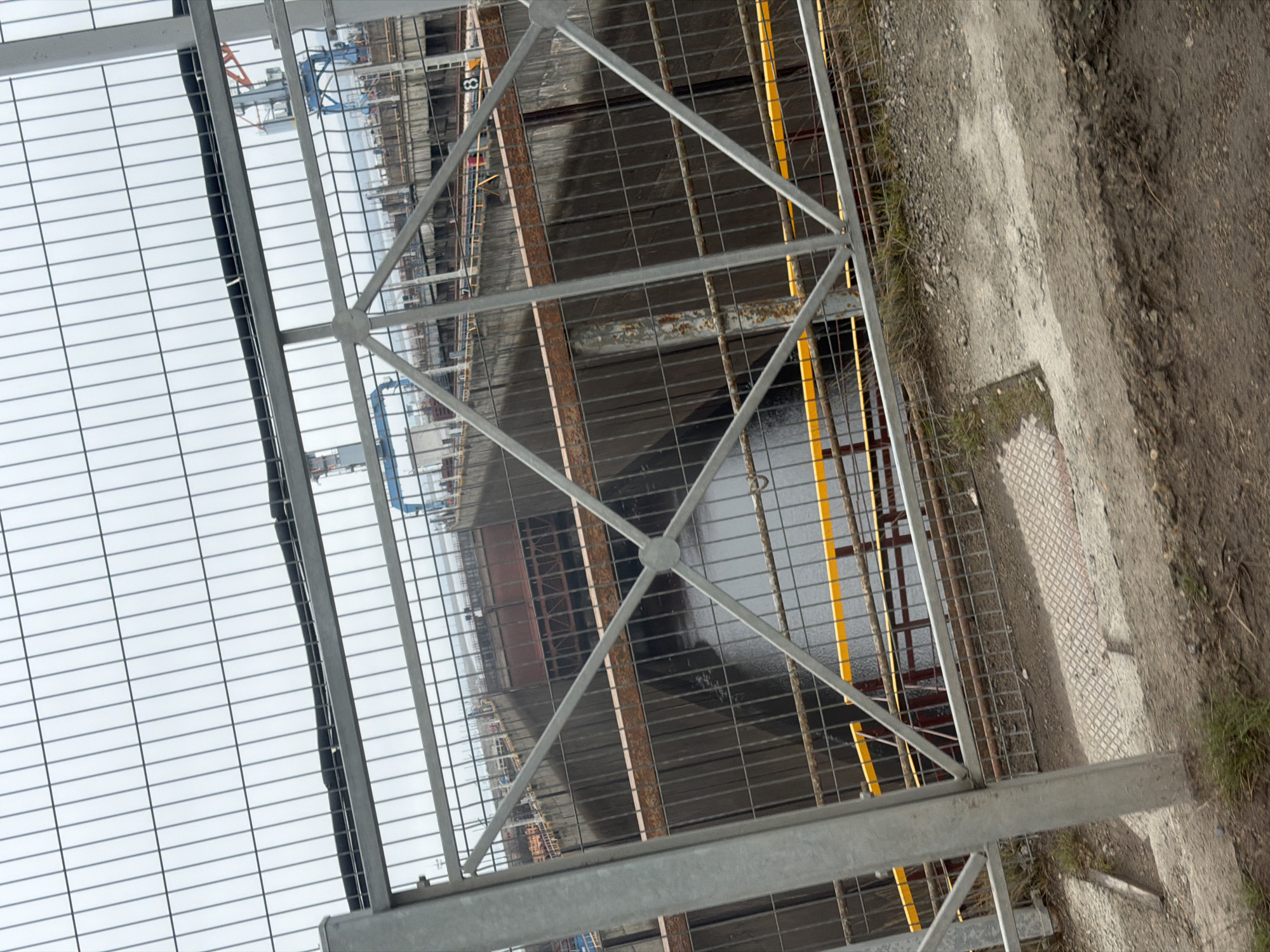 The image depicts a fenced-off area likely within an industrial or construction zone, possibly in Chaikovsky, Russia, given the user's specifications. The foreground is dominated by a gray, wire-mesh fence with structural metal supports. Beyond the fence, there is a pit or excavation filled with water, partially obscured by shadows. Orange or yellow structural elements are visible within the pit. In the background, there are industrial structures, cranes, and buildings under construction. The immediate area outside the fence is a dirt or gravel surface mixed with some vegetation. The sky is overcast, suggesting a cloudy day.