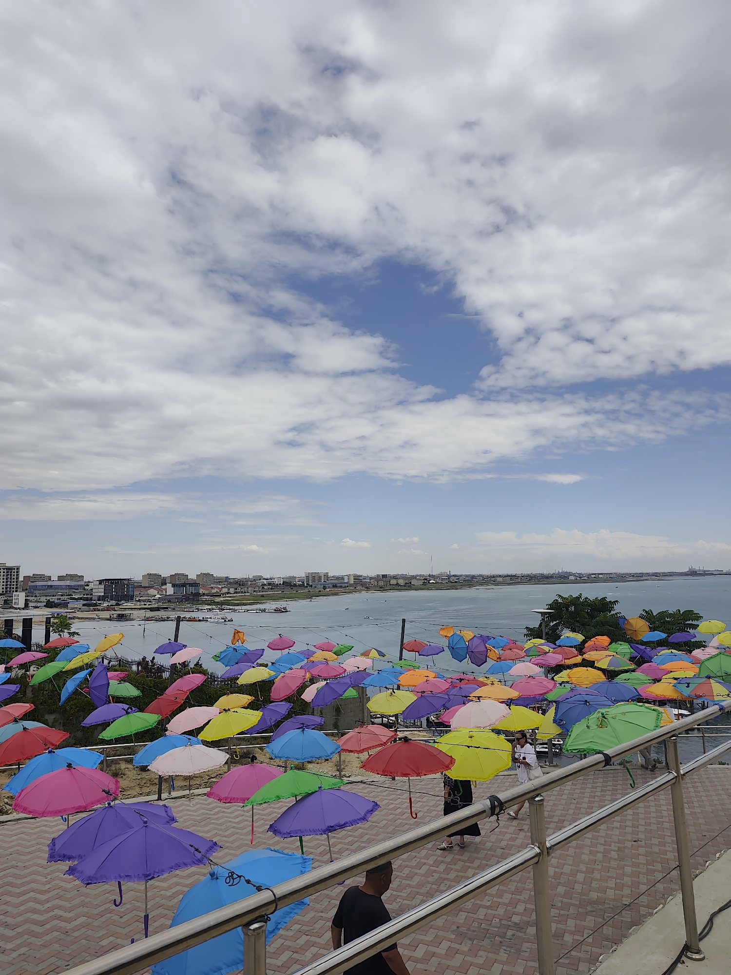 The image depicts a vibrant outdoor scene in Aktau, Kazakhstan, overlooking the Caspian Sea. A multitude of colorful umbrellas, in shades of pink, purple, blue, yellow, orange, green, and red, are suspended above a patterned walkway, creating a festive and artistic display. Some umbrellas are open, while others are closed. In the middle ground, a shoreline with buildings and a body of water, likely the Caspian Sea, is visible under a partly cloudy sky. A few boats are present on the water. Several individuals are walking on the walkway and viewing the umbrellas, some of whom are partially obscured. The walkway is paved with red and beige interlocking bricks and features metal railings. The overall impression is a lively public space, possibly a tourist attraction or a promenade, decorated with a unique umbrella installation.