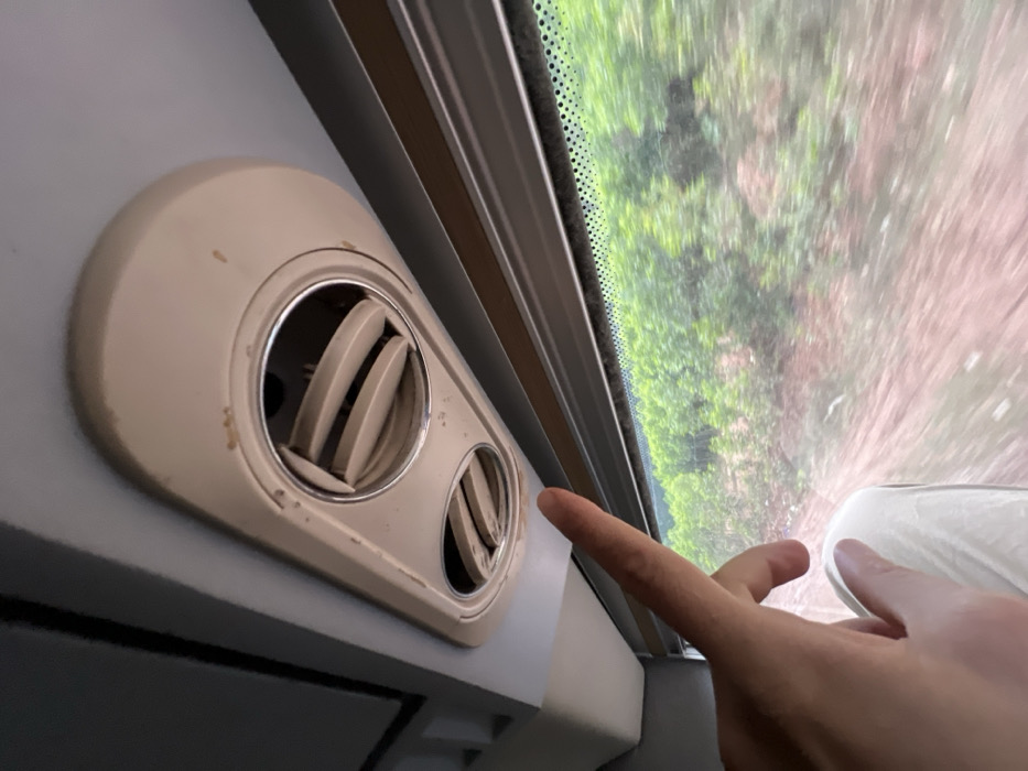 The image depicts the interior of a moving passenger vehicle, likely a bus or train, within or en route to Aê Thi, Vietnam. A light-skinned human hand, positioned in the lower-left quadrant, extends its index finger upwards, pointing towards a cream-colored overhead fixture. This fixture contains two circular air vents with visible adjustable slats, one larger than the other. The fixture exhibits visible wear, scuff marks, and dirt accumulation. Below the overhead fixture and the pointing hand, a window reveals a blurred exterior landscape, characterized by green foliage and reddish-brown soil, consistent with motion. Part of a person's leg or lap, covered in light-colored fabric, is visible in the lower-left.