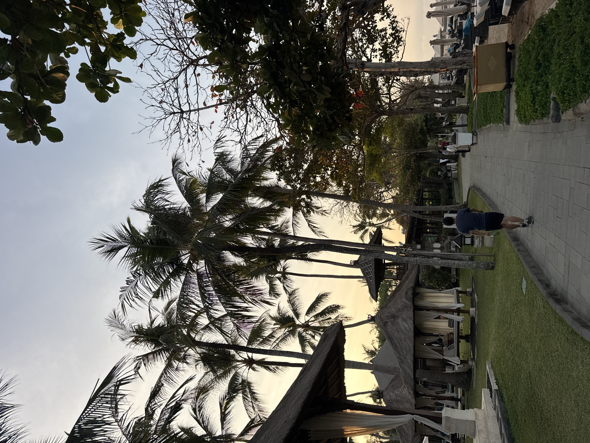 This image depicts a resort scene in Kuta Selatan, Indonesia, likely during the late afternoon or early evening. The foreground features a paved walkway leading away from the viewer towards a line of thatched-roof cabanas with white curtains. A lone man in a dark blue t-shirt and black shorts is walking away from the camera on the walkway. Lush green grass surrounds the cabanas, and numerous tall palm trees are scattered throughout the scene, some with their fronds silhouetted against the pale sky. In the background, more trees and structures are visible, and beyond that, beach chairs and umbrellas suggest proximity to a beach. The lighting indicates a soft, diffused natural light, typical of twilight. The overall atmosphere is serene and suggests a tropical vacation setting.