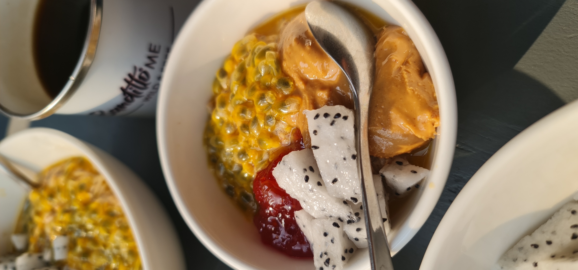 A high-angle, close-up shot depicts two white bowls and a metal mug on a dark, textured surface. The main focus is a bowl filled with passion fruit pulp, two scoops of peanut butter-colored ice cream, chunks of dragon fruit, and a dollop of reddish jam. A spoon rests in the bowl. Part of another bowl containing a similar fruit mixture is visible to the left. The metal mug, holding a dark liquid, is visible at the upper left of the frame.
There is no indication this is Đà Nẵng, Vietnam.