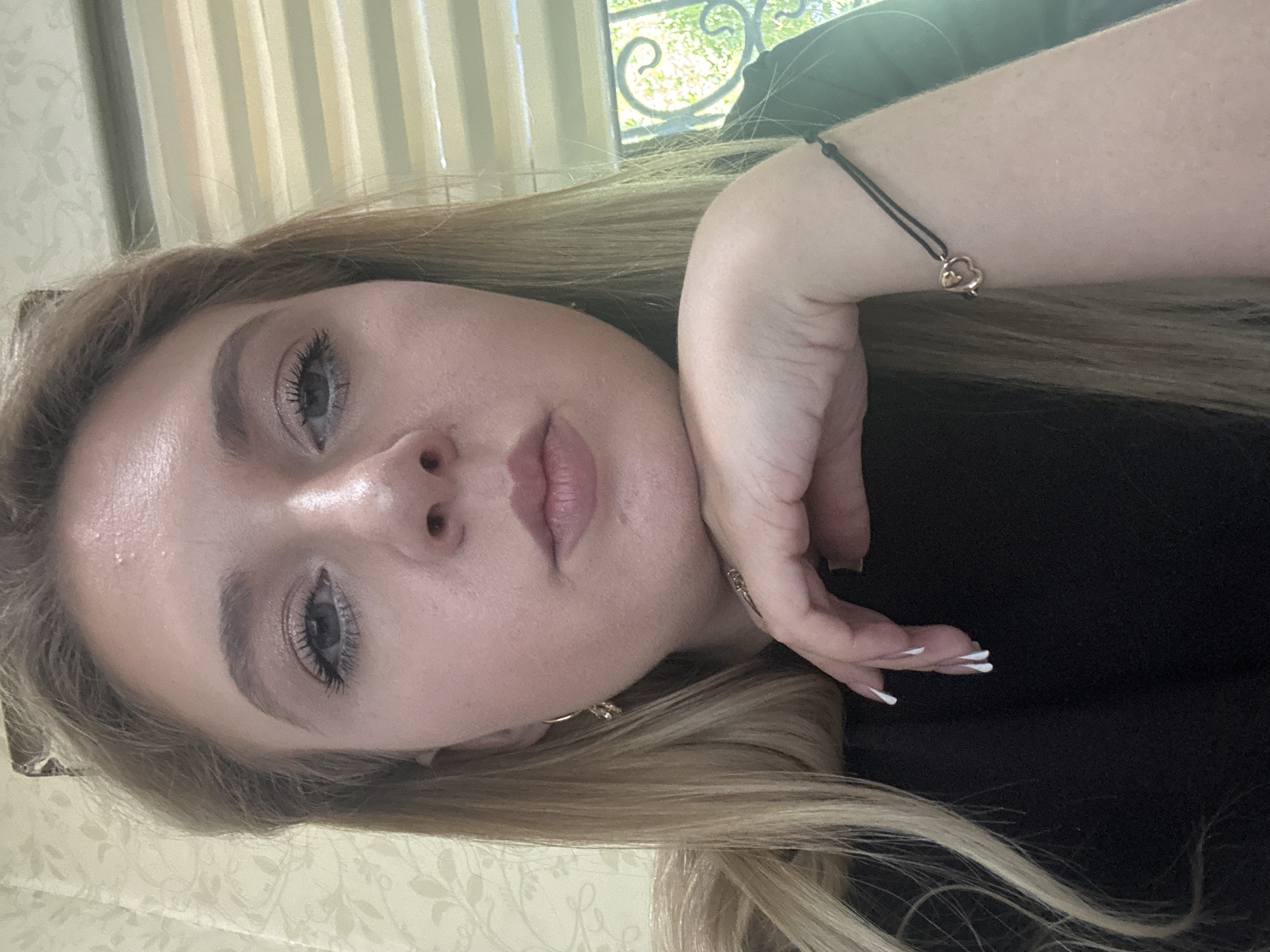 A young woman with blonde hair is looking directly at the camera, resting her chin on her hand. She is wearing a black top and has a bracelet on her wrist with a heart-shaped charm. Her nails are painted white. The background is a wall with a floral pattern and blinds.