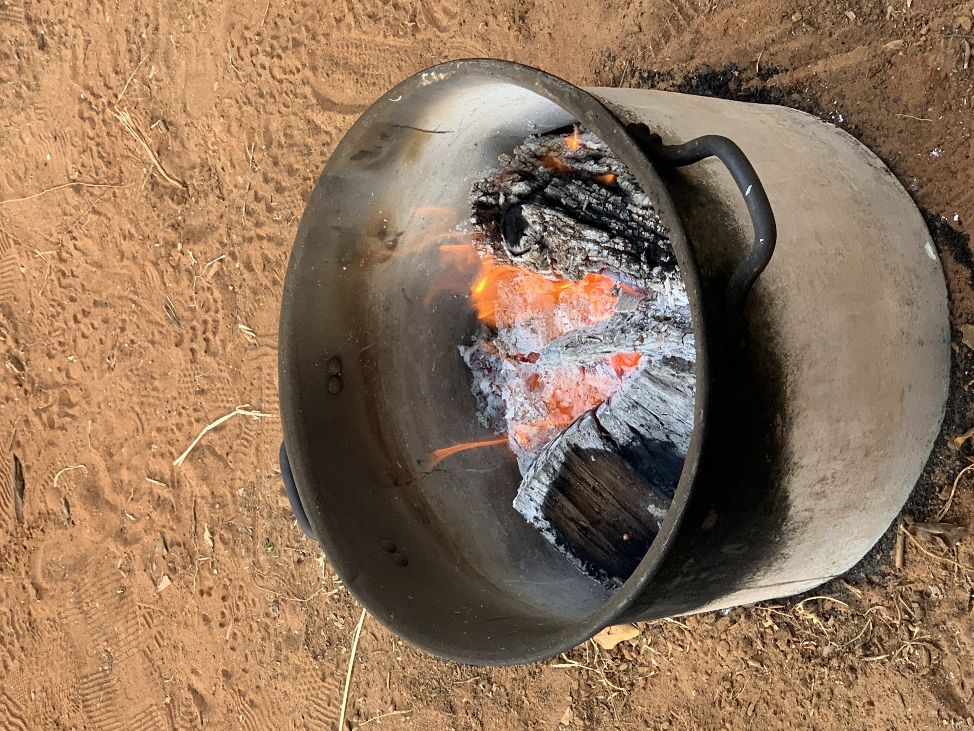 The image shows a metal fire pit on reddish-brown dirt, likely outdoors. Inside the pit, there are burning logs with visible flames and white ash. The fire pit is cylindrical with a handle on one side. The surrounding dirt shows signs of being disturbed with footprints and small pieces of organic material. The image does not contain enough information to determine if it was taken in Gan Sorek, Israel.