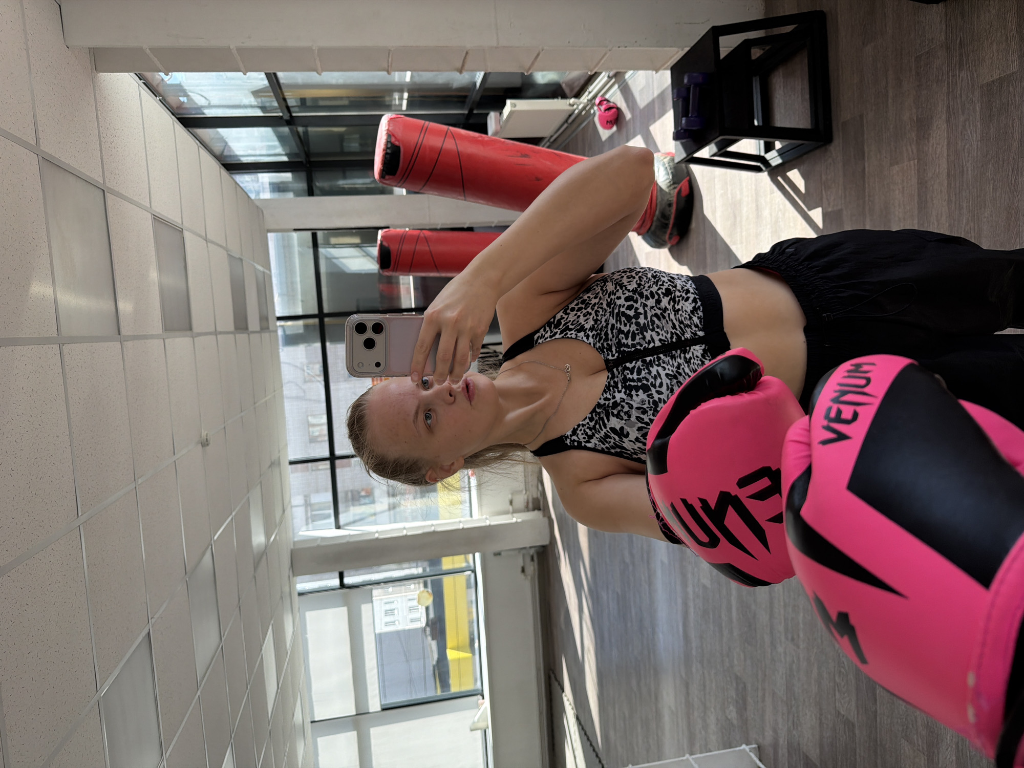 A woman in a black and white sports bra, black pants, and pink boxing gloves takes a selfie with an iPhone in a gym. She stands near a window with a view of buildings in Izhevsk, Russia. The gym has red punching bags, a black shelf, and dumbbells.