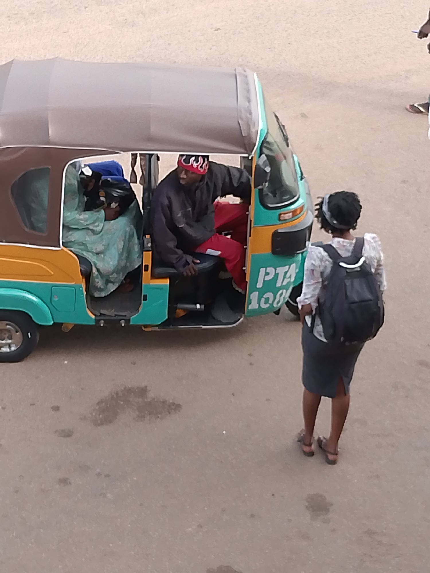 In the city of Bukuru, Nigeria, a man wearing a red bandana sits in the driver's seat of a teal and yellow auto-rickshaw. A passenger, dressed in a teal garment, is seated behind him. The auto-rickshaw is parked on a paved surface, which appears to be a street, with some scattered dark spots. A woman wearing a white shirt with a floral pattern and a dark gray skirt stands facing the auto-rickshaw. She has a dark backpack on her back and is wearing sandals. Text on the side of the auto-rickshaw is partially visible, with "PTA" and "100" clearly legible. The overall scene suggests a moment of transition, possibly a stop for passengers or a brief interaction.