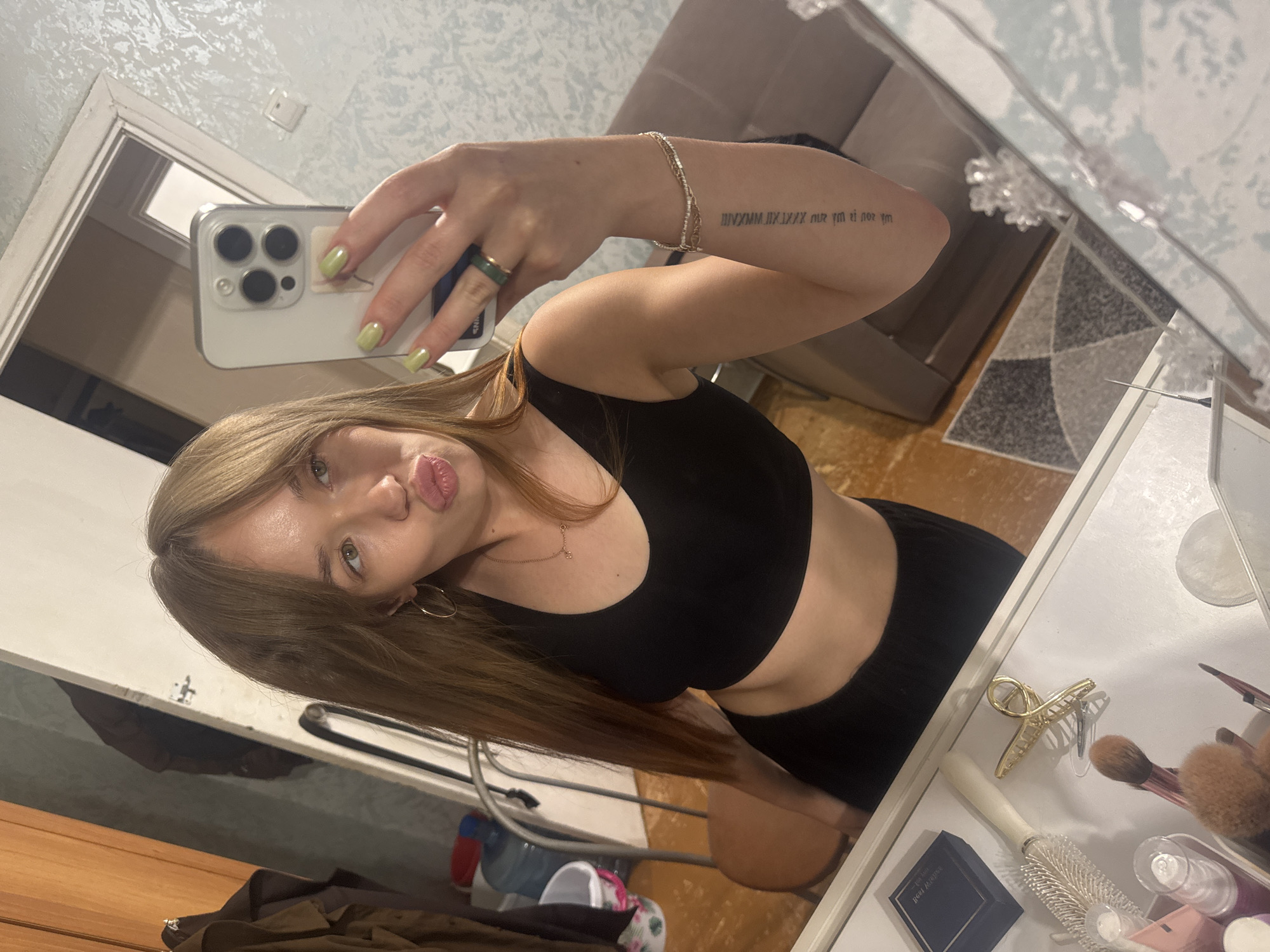 A young woman with long, straight blonde hair is taking a mirror selfie in what appears to be a bedroom or dressing area. She is wearing a black crop top and black bottoms, exposing her midriff. Her right hand holds a white smartphone, and her left arm is bent, showing a tattoo on her forearm and a bracelet. She has a green ring on her right index finger and light green nail polish. Her lips are pursed, and she is looking at the phone's screen. On the vanity in front of her, there are various makeup brushes, a hairbrush, a blue jewelry box, and a gold hair clip. In the background, there is a closet with clothes, a chair, a water cooler, and a patterned rug on the floor. The wallpaper appears to be a light blue textured pattern. The scene does not contain any elements that would indicate it is located in the city of Chaikovsky, Russia, or any specific urban location.