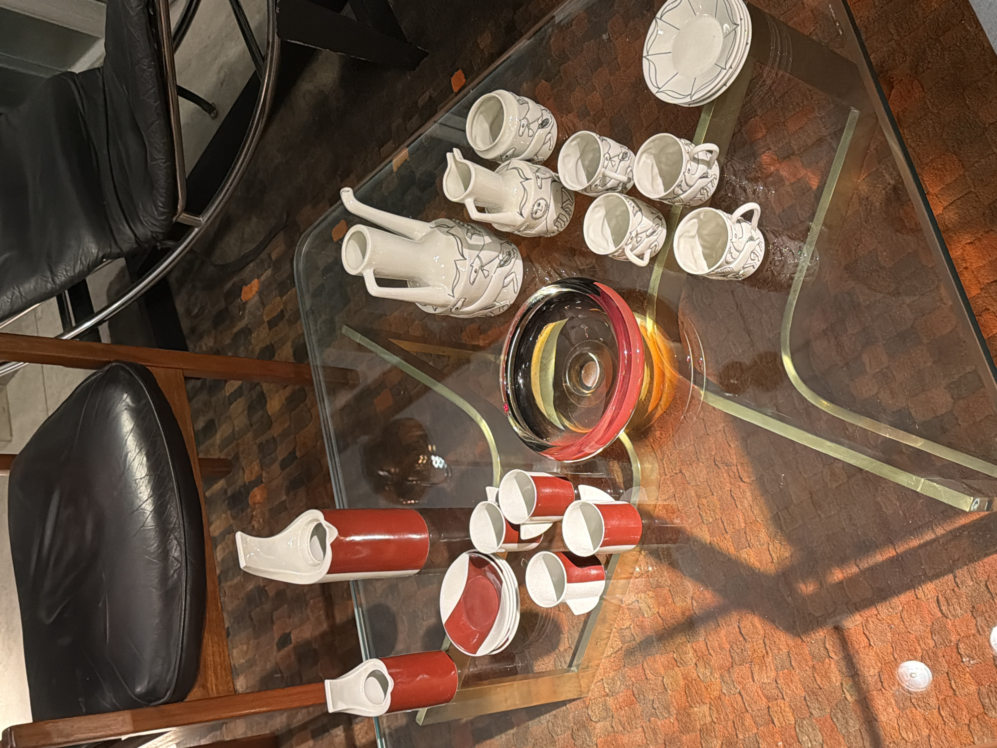 A high-angle shot displays a glass-topped table. Two chairs with black leather seating are positioned on either side. The tabletop exhibits two sets of dishware. The first set features white dishes with black geometric patterns, comprising plates, a pair of pitchers, and a collection of cups. The second set presents a red and white color scheme, including cups, saucers, and pitchers. A transparent glass bowl with red and amber accents is also visible. The setting is an interior, possibly a room within a building. The location is Buenos Aires, Argentina. The table is set with objects, but no active human presence or interactions are evident. The environment is a well-lit, indoor space.